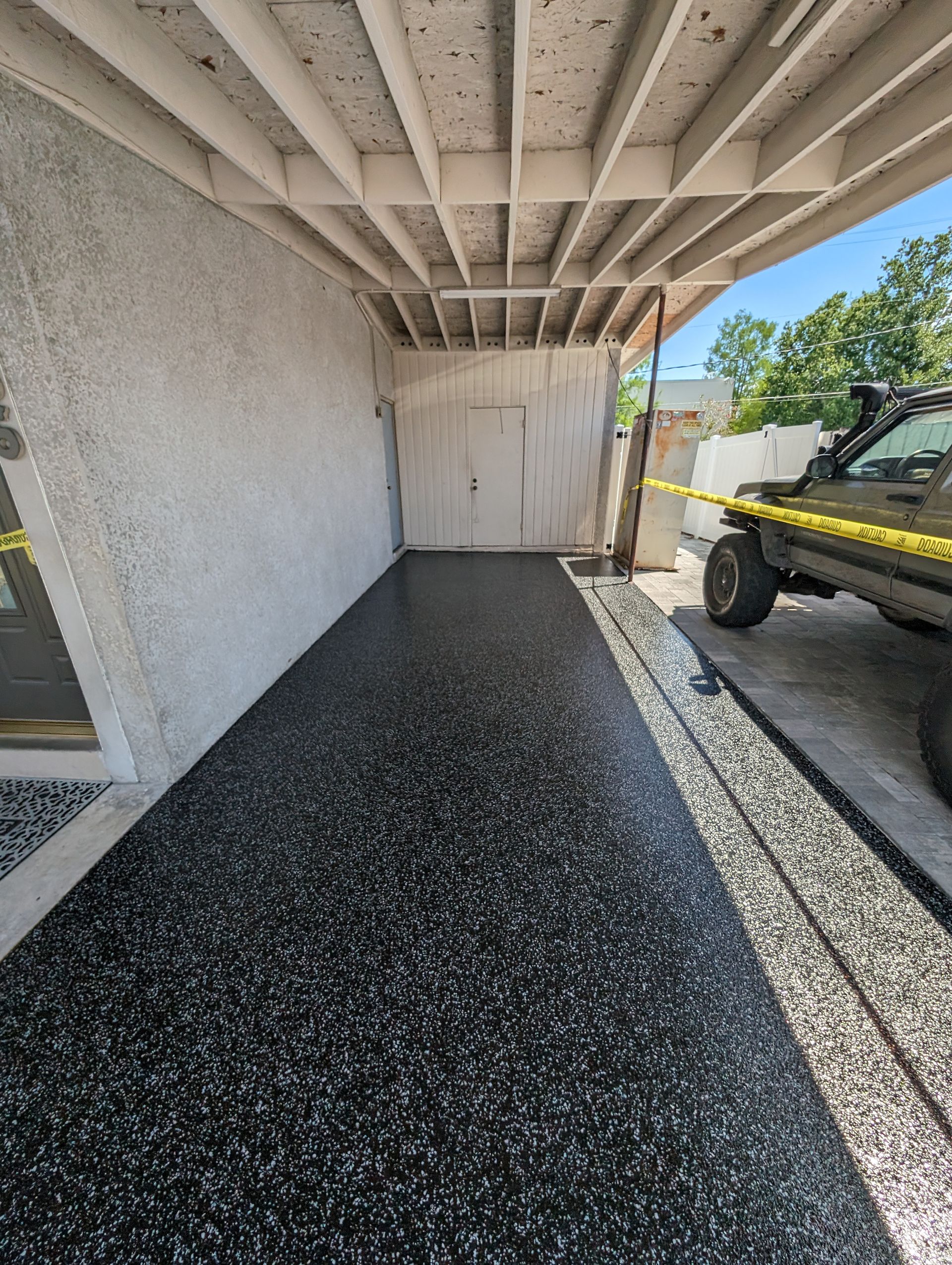 Covered driveway beside a stucco house, with black resurfaced floor and a utility trailer parked on the right.