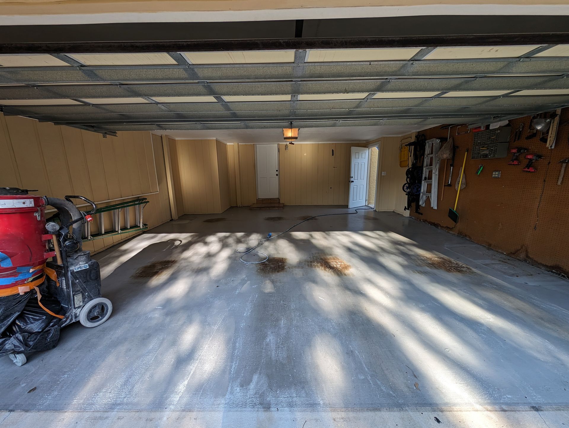 Empty garage with concrete floor, open door, shelves and tools along the walls, sunlight casting shadows.