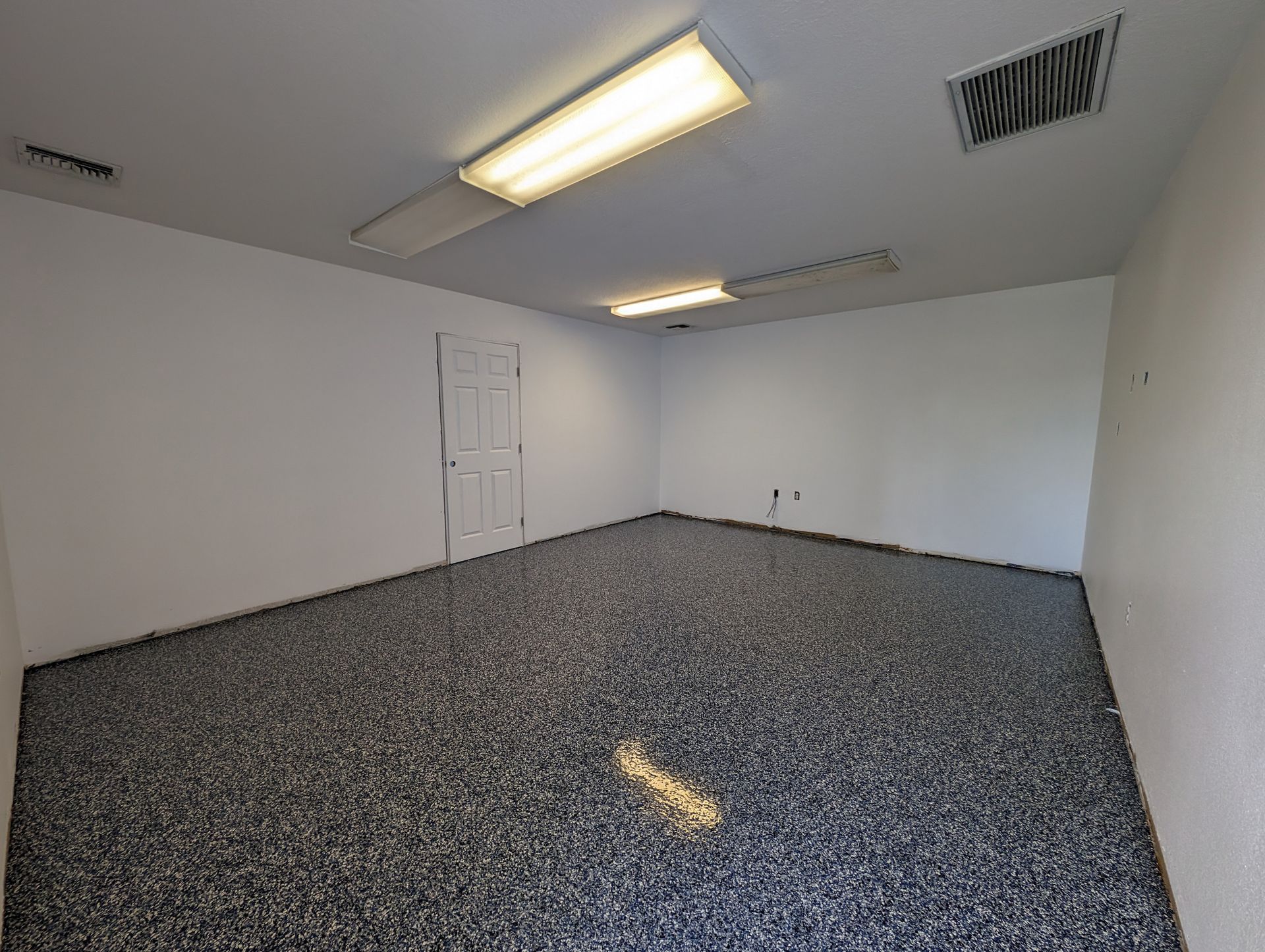 Empty office room with white walls, speckled floor, fluorescent ceiling lights, and a door.
