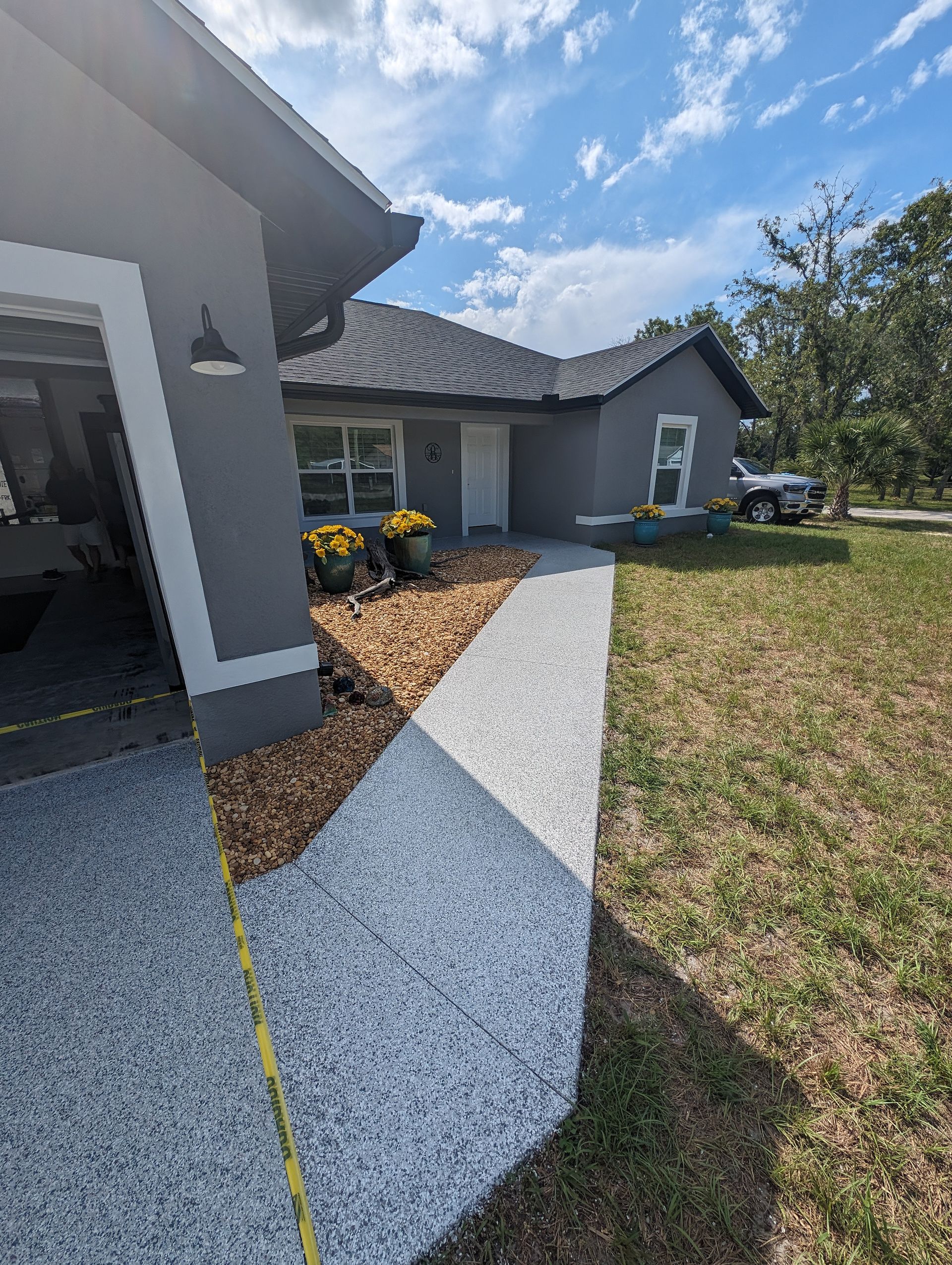 Front porch walkway with white gravel, dark roof, yellow flowers, and a grassy yard under a bright blue sky.