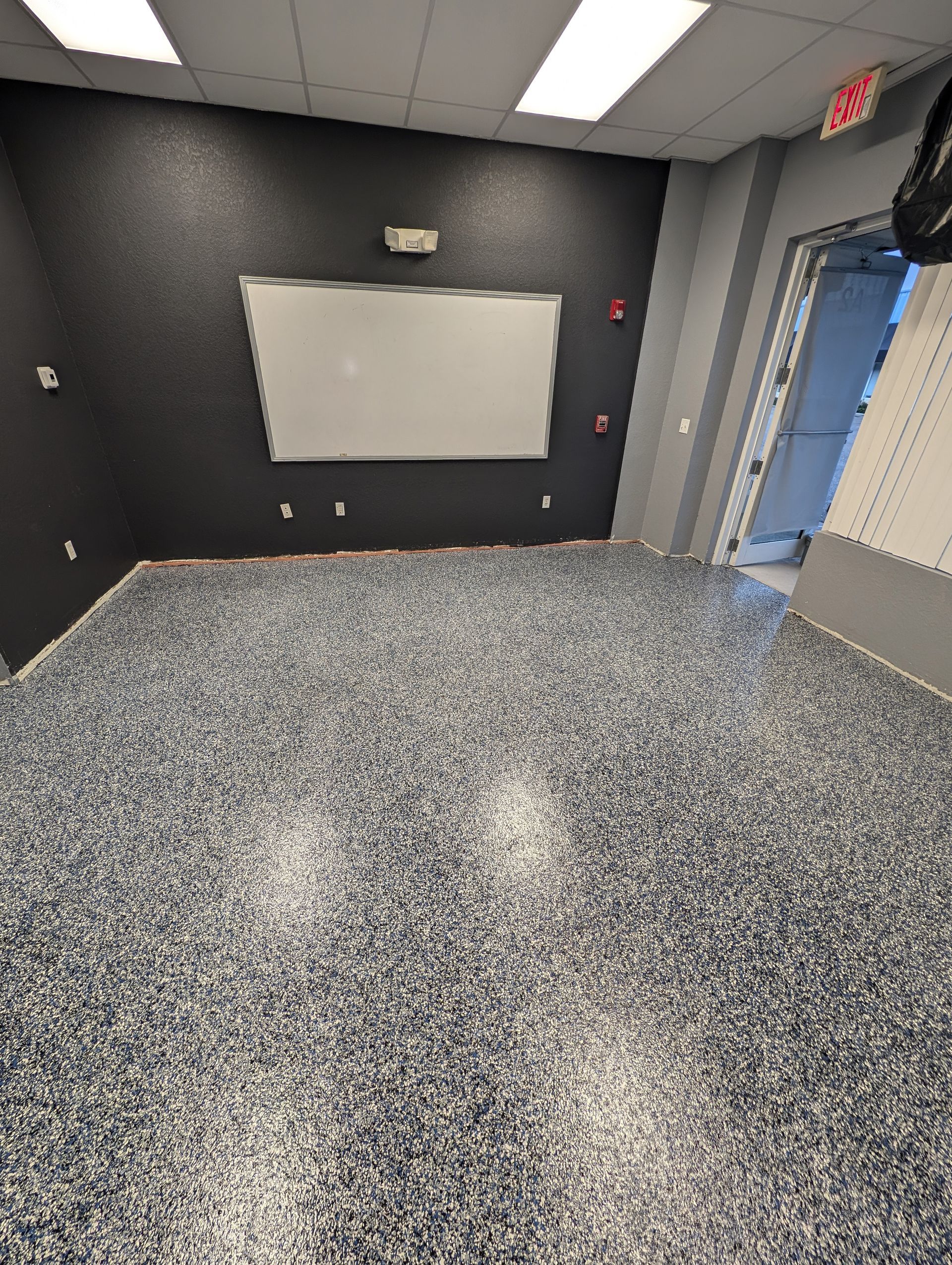 Empty black room with speckled floor, wall-mounted whiteboard, and open door to a blue-lit hallway