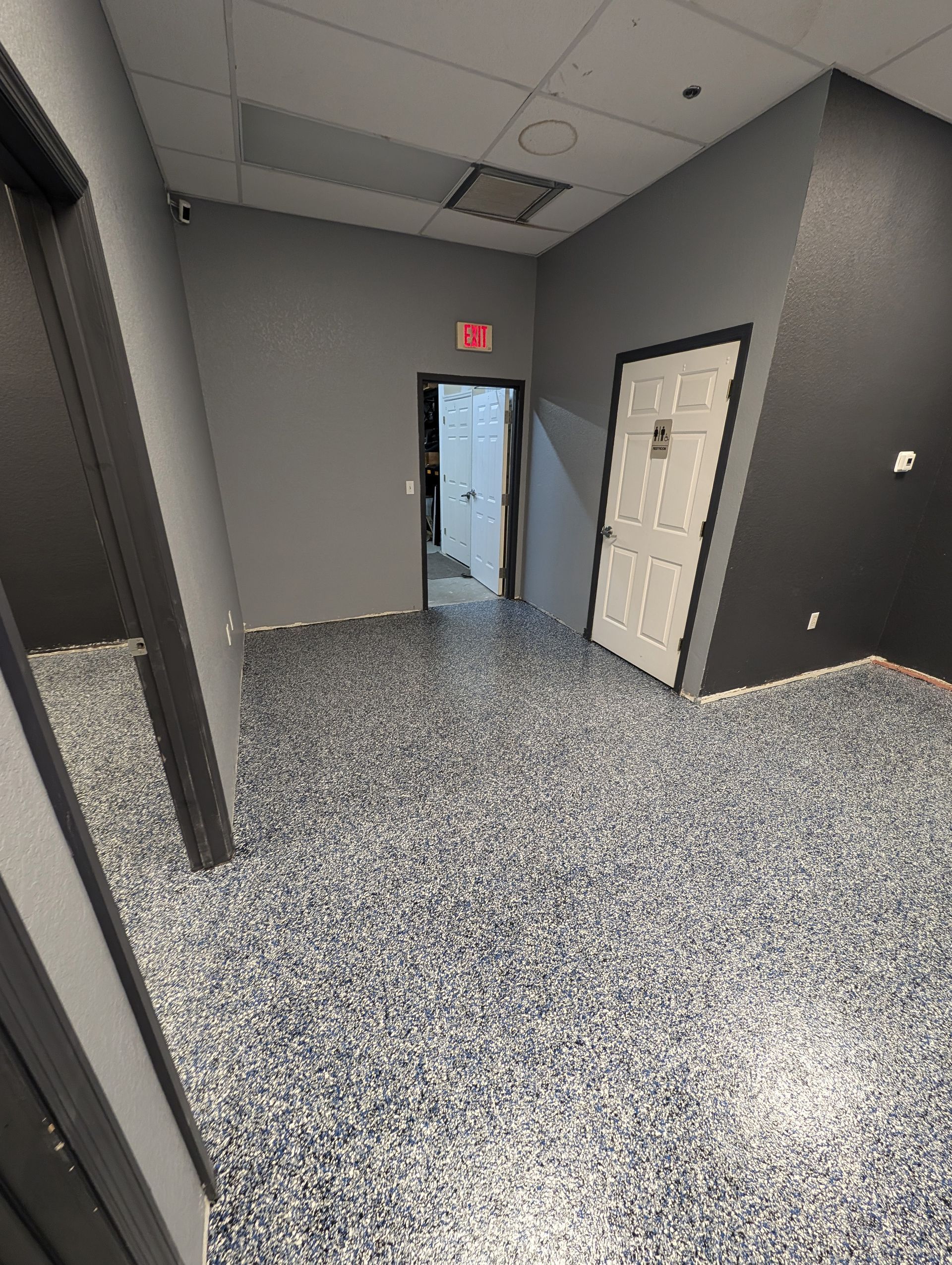Empty gray hallway with speckled carpet, closed doors, and an exit door with red sign.