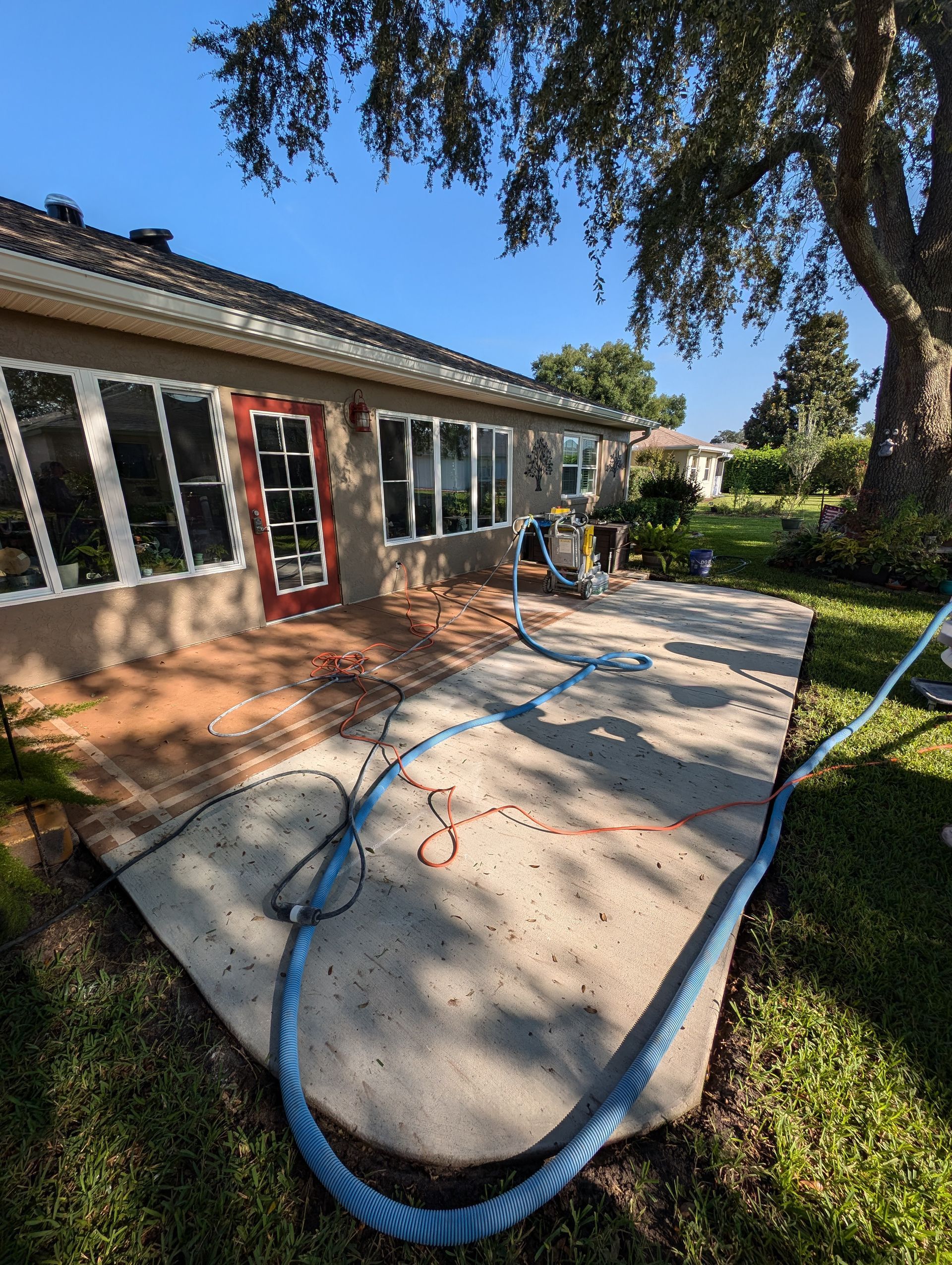 House patio under construction with blue hoses, red extension cords, and a concrete slab beside a garden