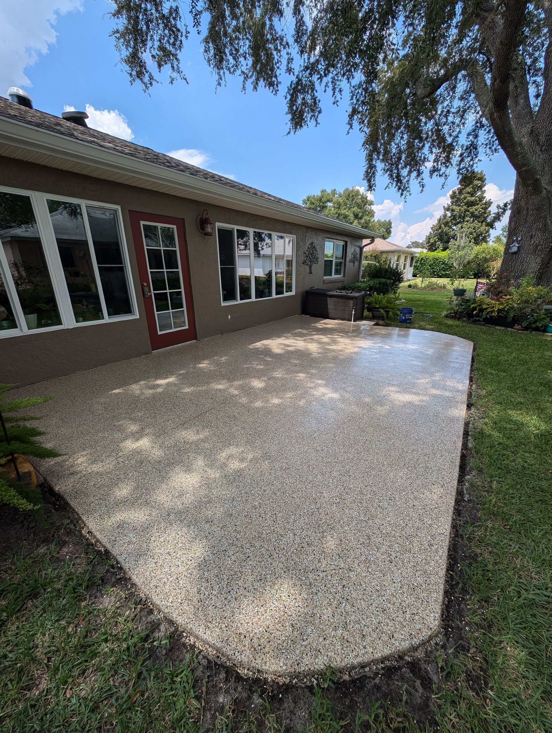 Backyard patio with light gravel surface beside a house, bordered by grass and trees.
