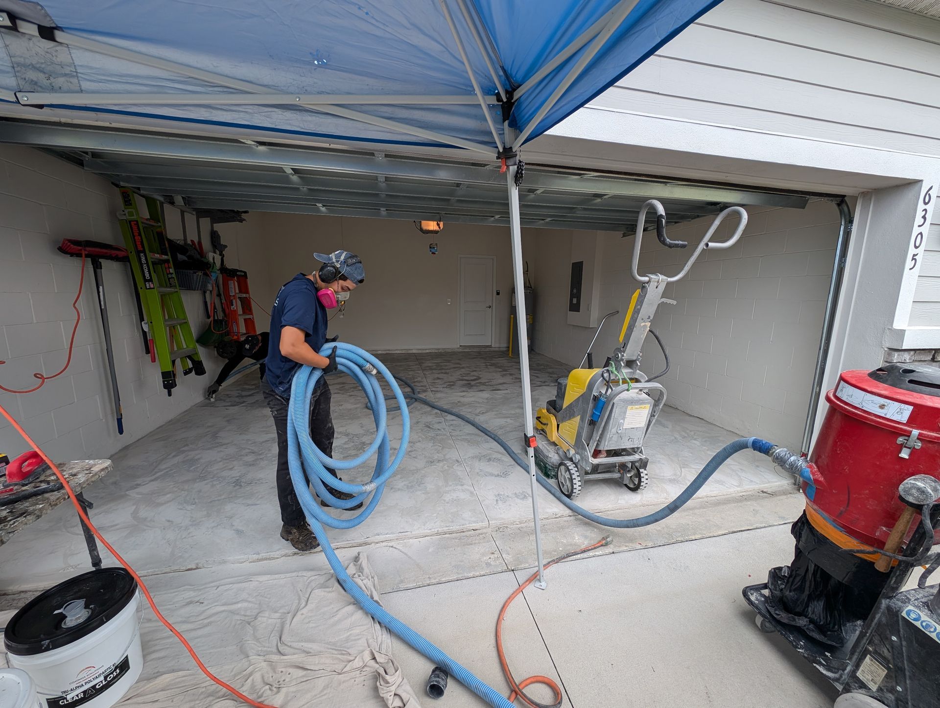 Worker coiling blue hose in garage under canopy, with floor equipment and tools nearby.