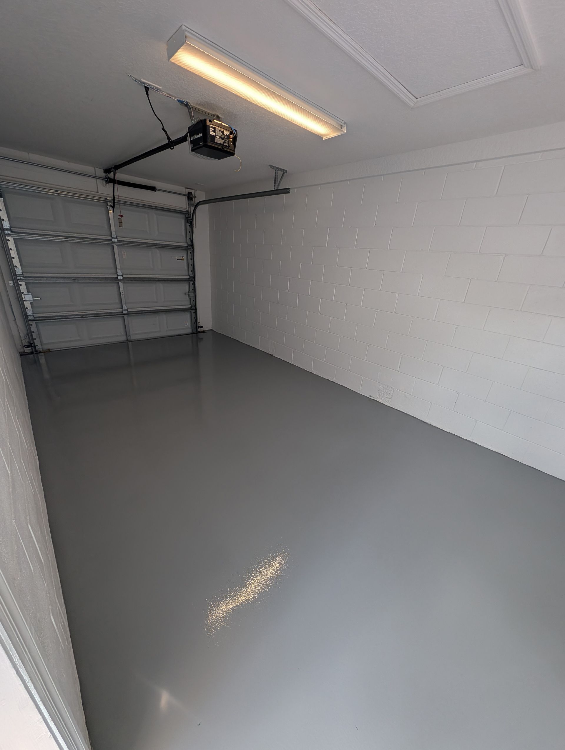 Empty garage with gray floor, white walls, shelving on the left, and an overhead light.