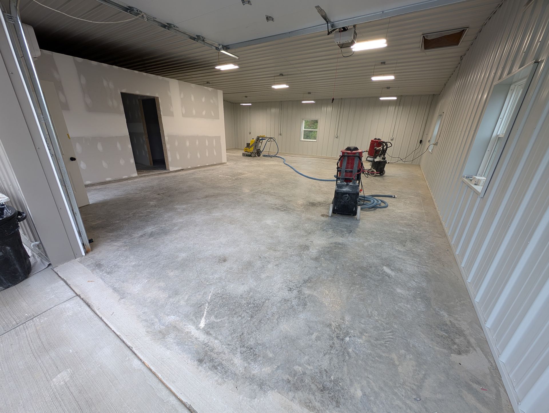 Empty warehouse room under renovation with bare concrete floor, ceiling lights, and two floor machines.