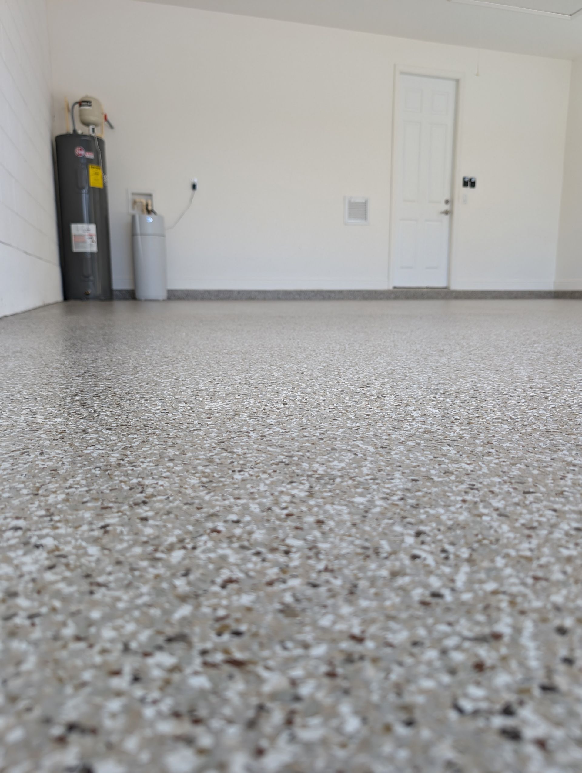 Empty garage with speckled concrete floor, white walls, a water heater, and a closed white door.