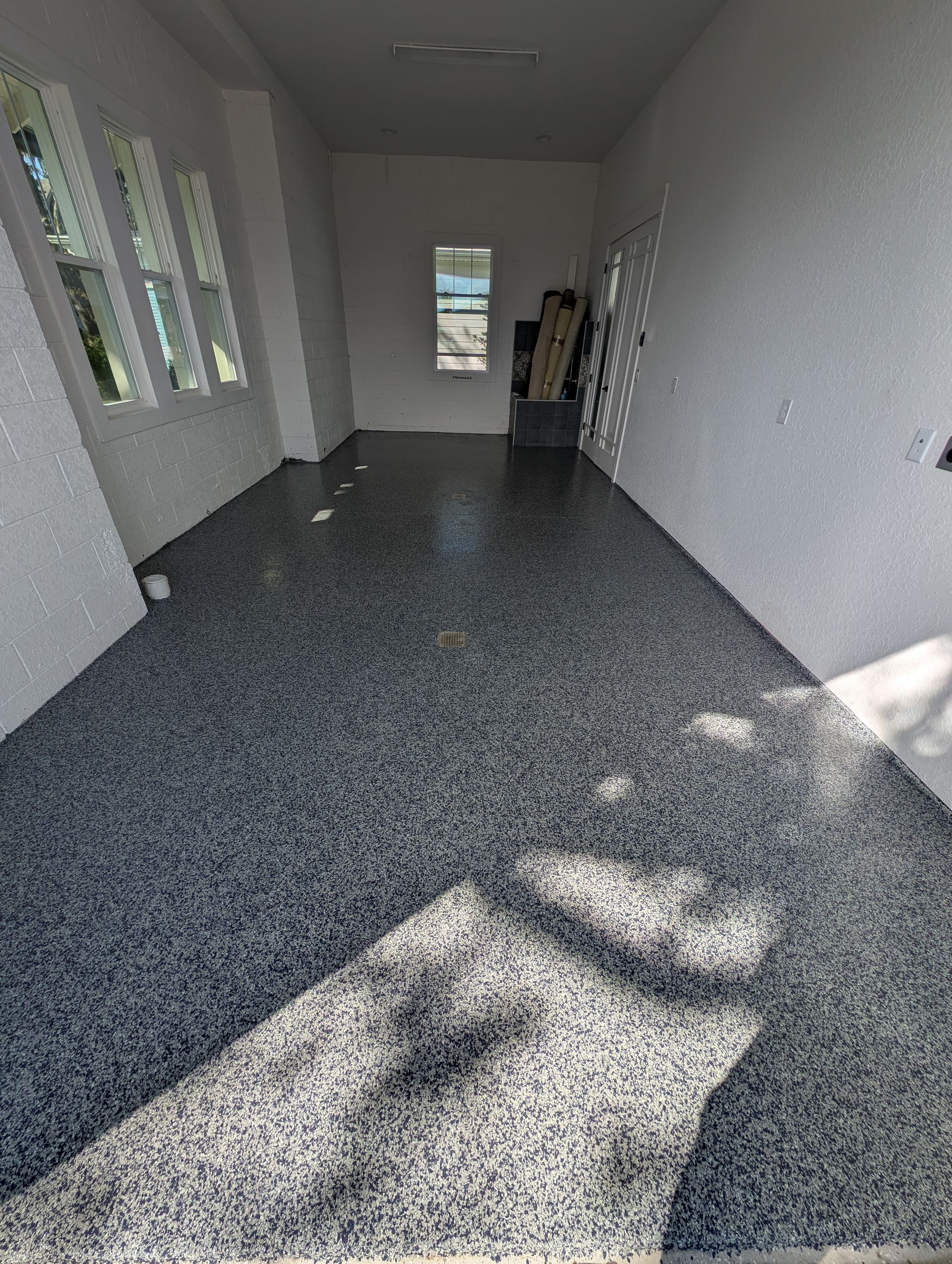 Empty narrow room with white walls, gray speckled floor, windows on the left, and a person standing at the far end.