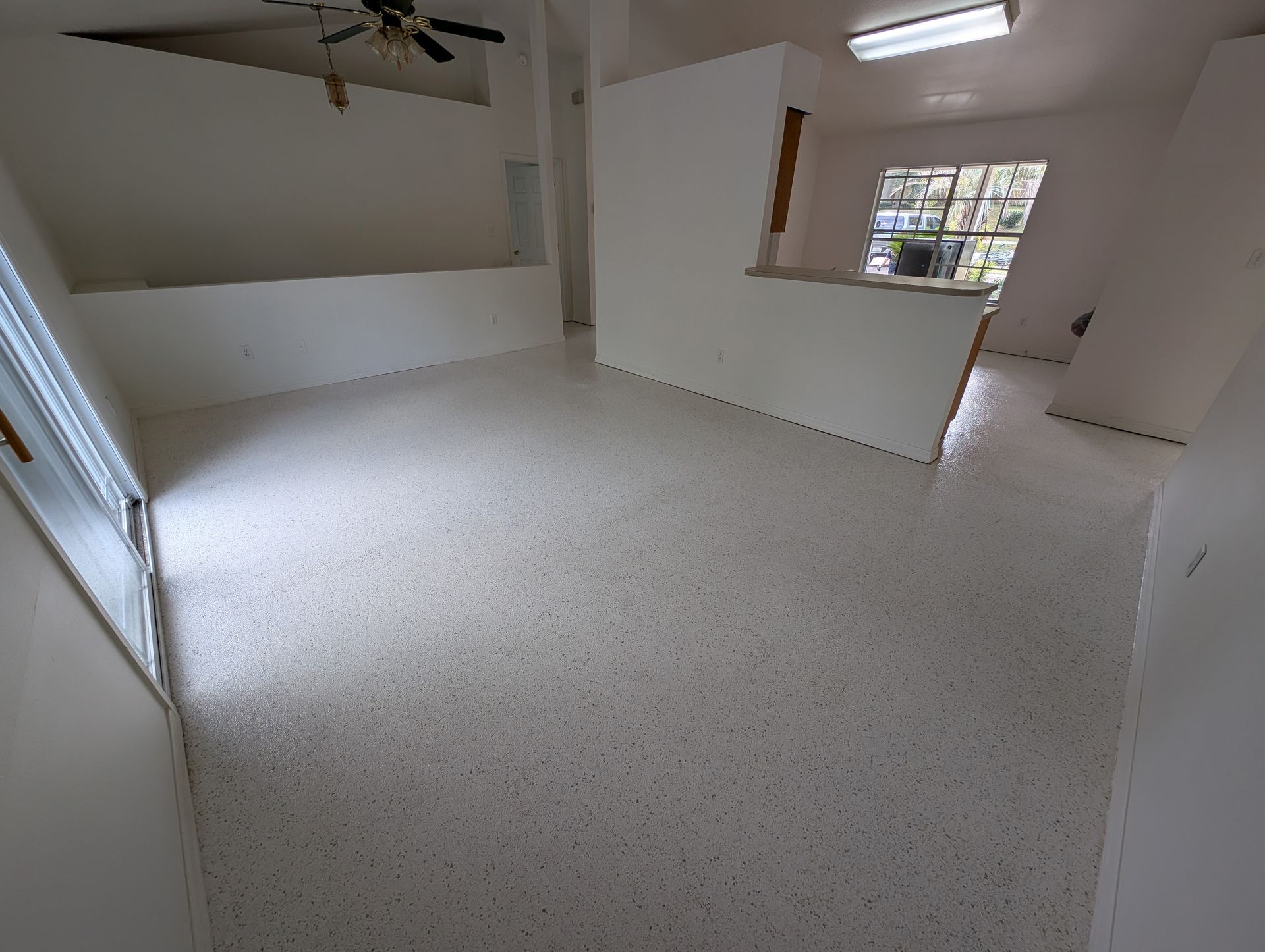 Empty white room with speckled floor, ceiling fan, and a small window opening to another room