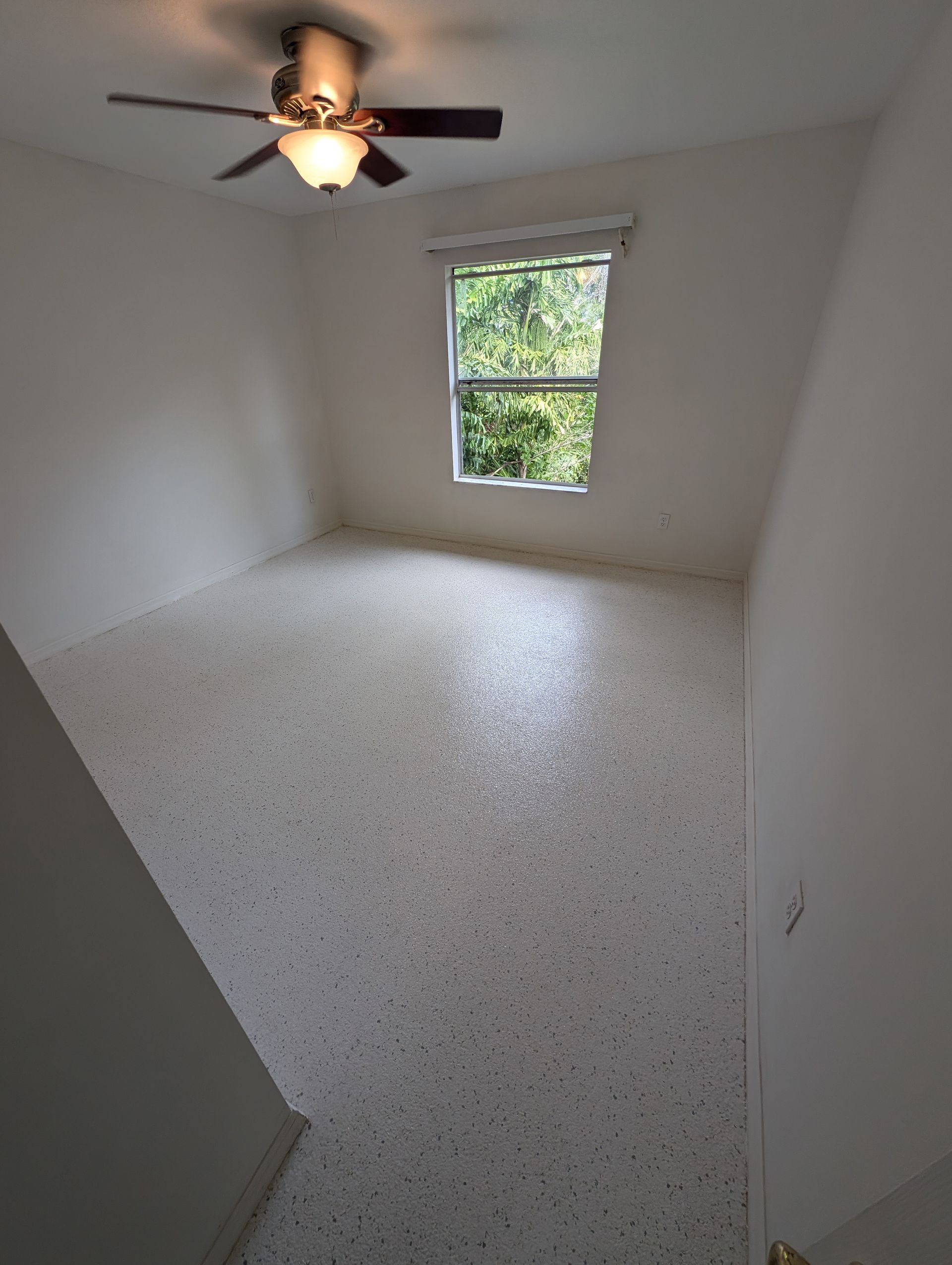 Empty small bedroom with white walls, speckled floor, ceiling fan, and one window.