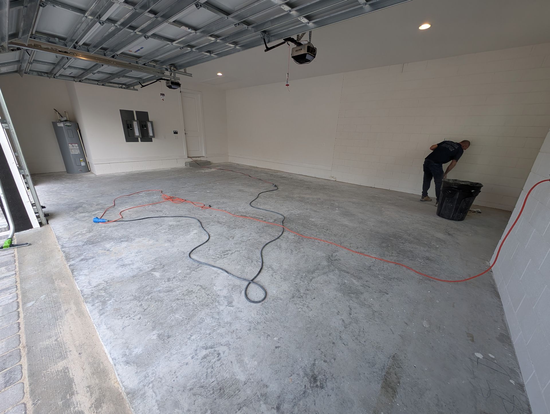 Empty garage under renovation, with a worker near the back wall and cords across the concrete floor.
