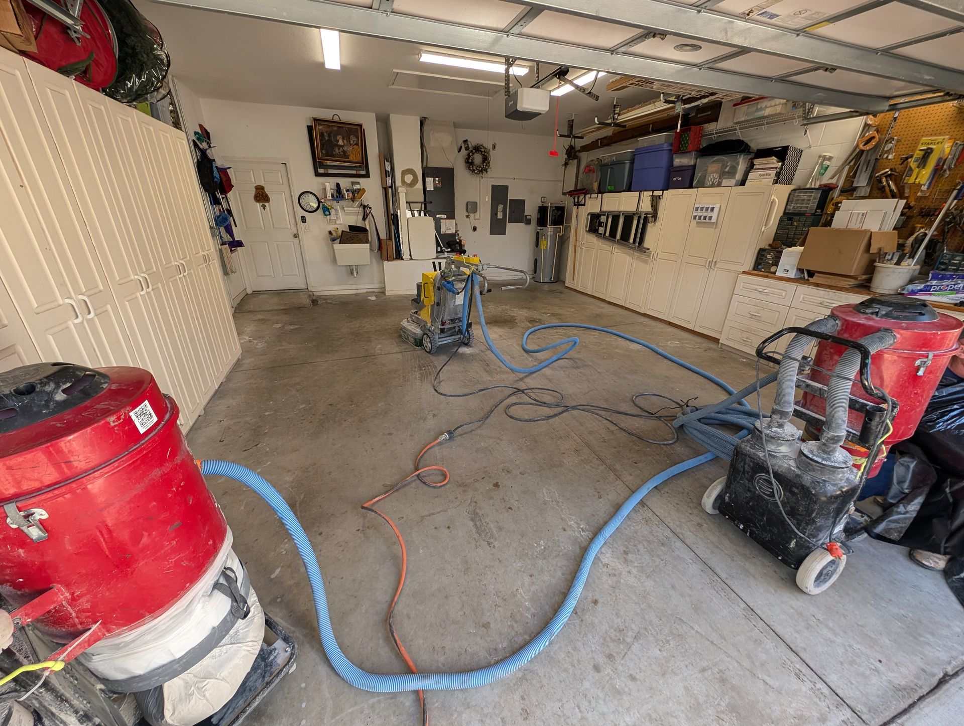 Garage with concrete floor, red and yellow industrial floor-grinding machines, and blue hoses laid across the floor.