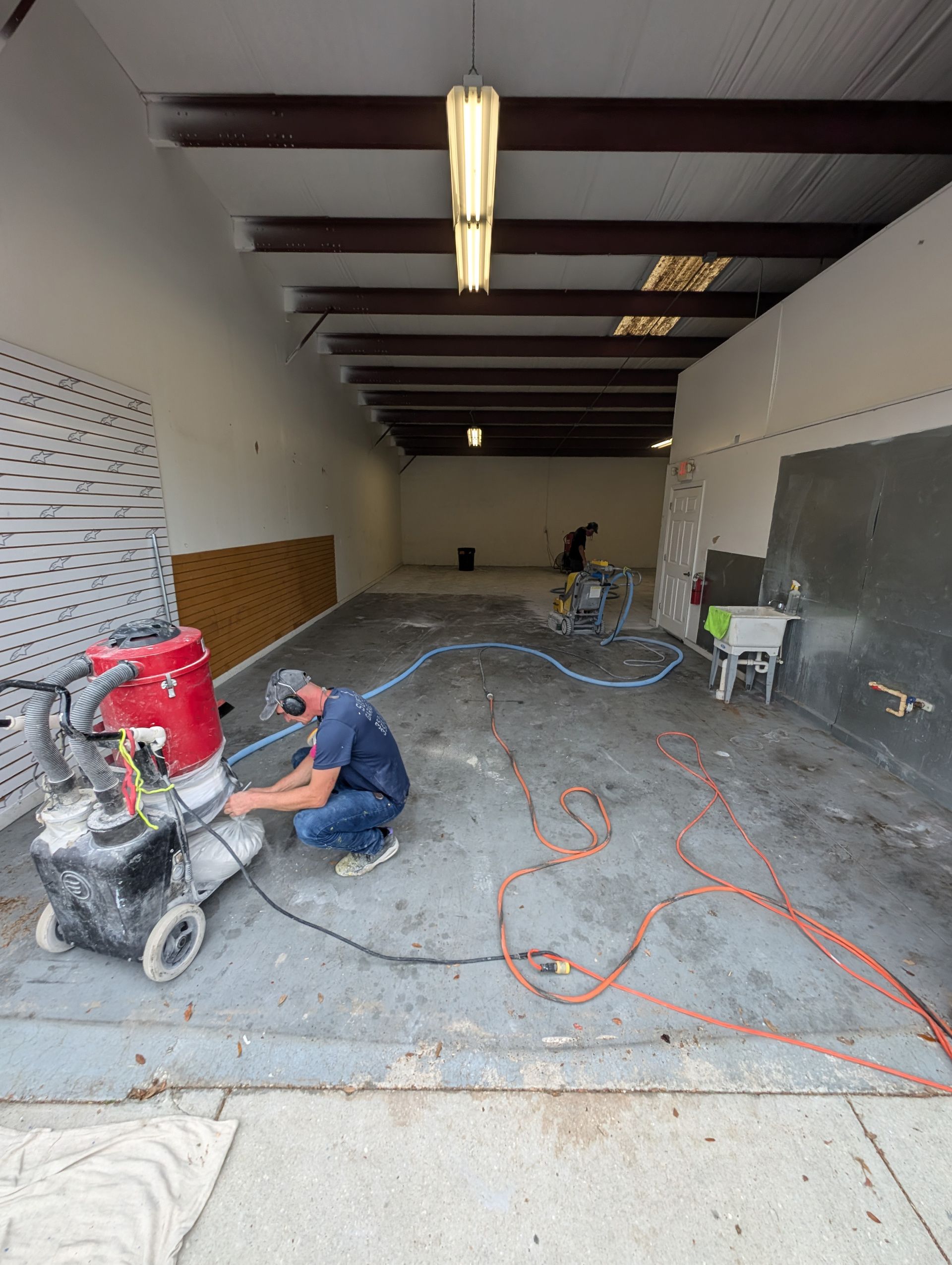 Workers sanding a concrete floor inside a long, unfinished warehouse space
