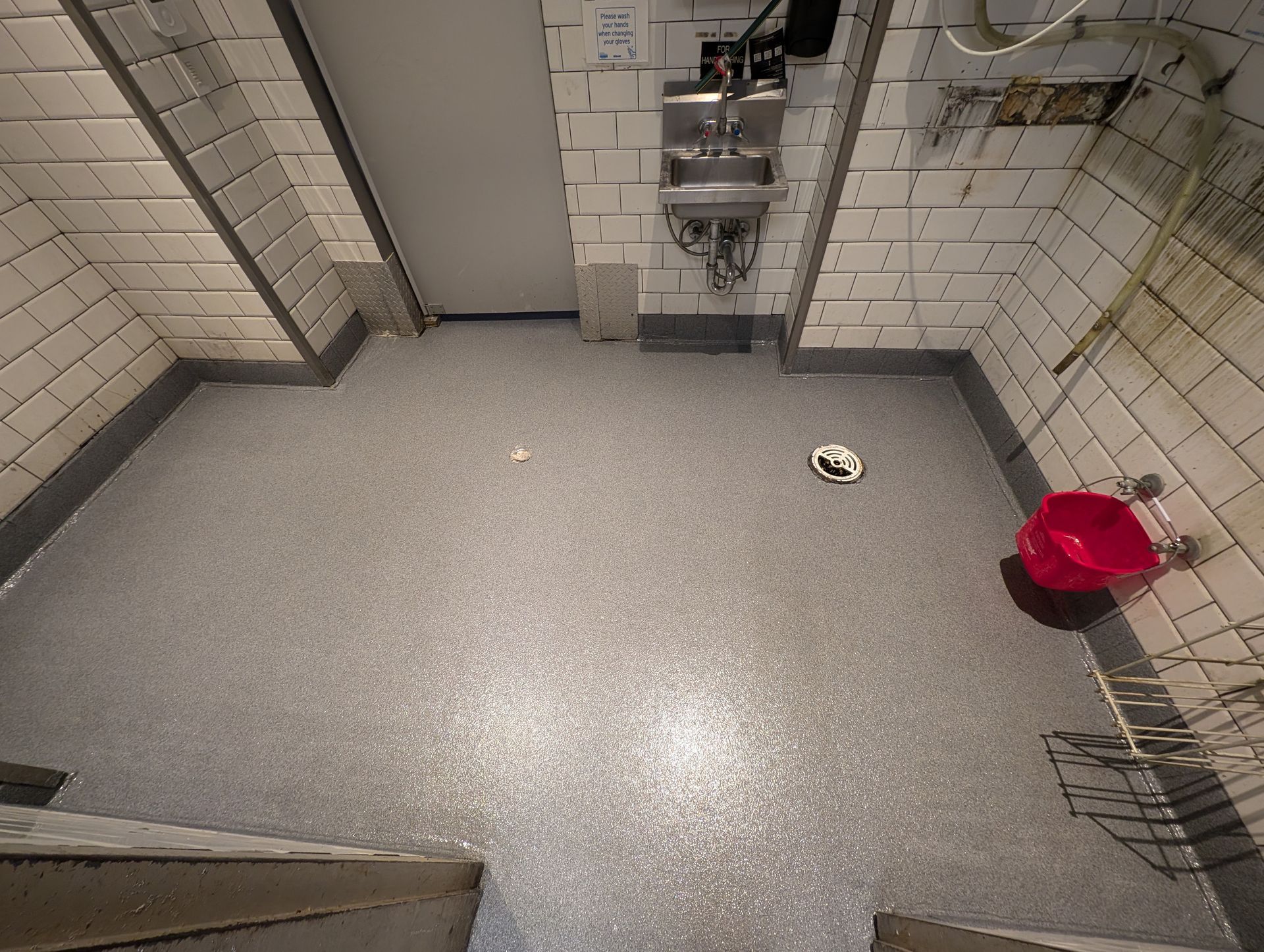 Empty tiled restroom with a gray floor, metal fixtures, and a red trash bin in the corner