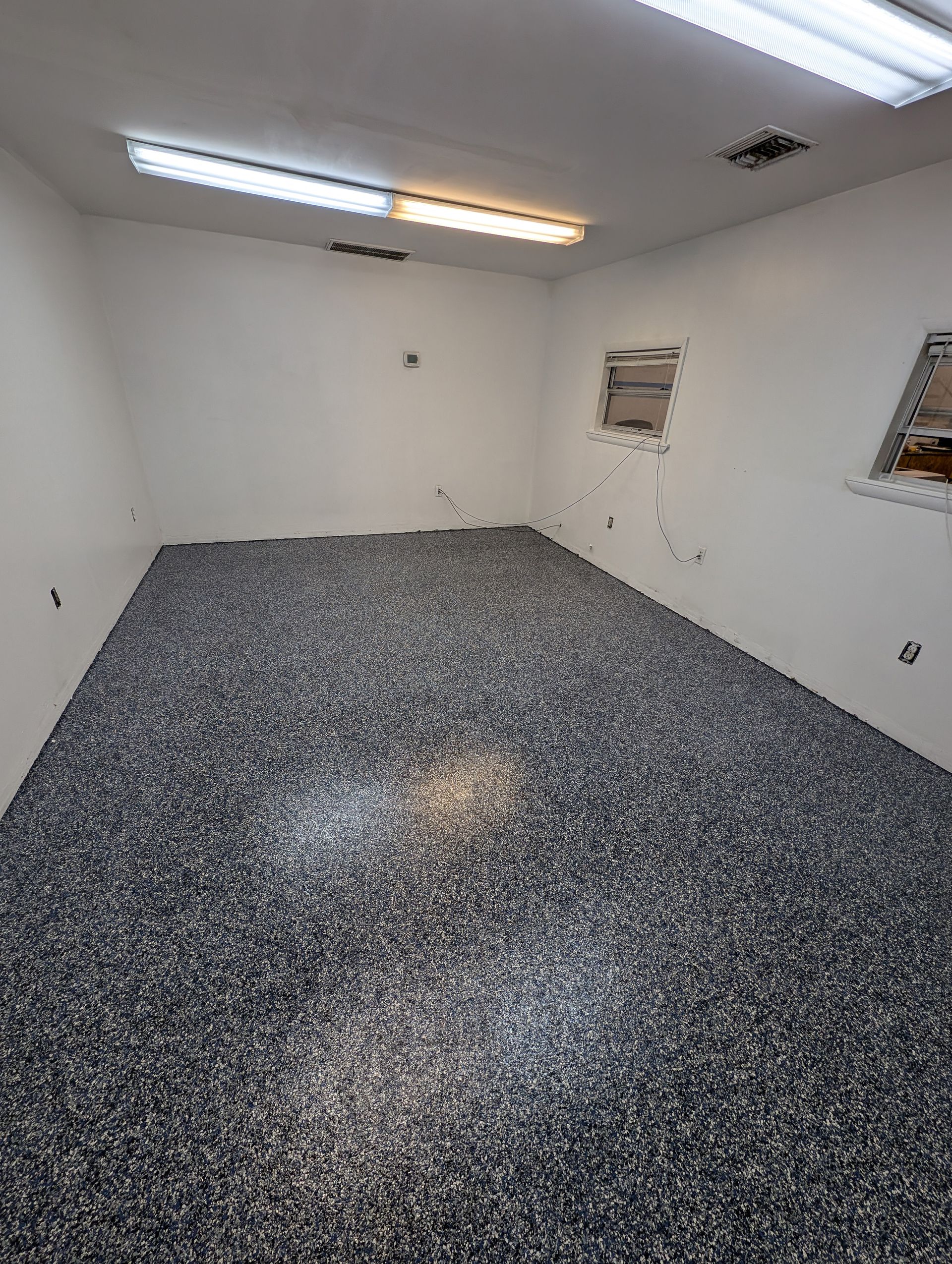 Small white room with speckled gray floor, fluorescent ceiling lights, and two wall vents.