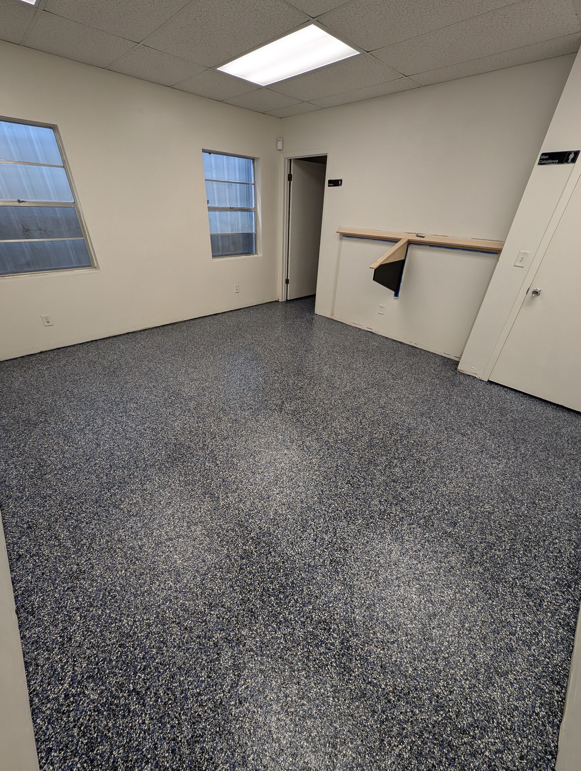 Empty room with gray carpet, beige walls, two windows, and a wall shelf near an open doorway.