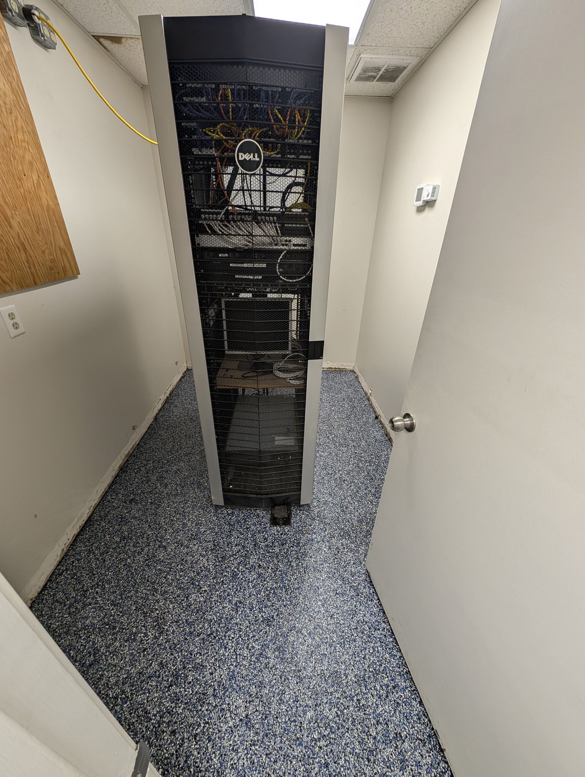 Server rack in a narrow utility room with exposed cables and speckled flooring.
