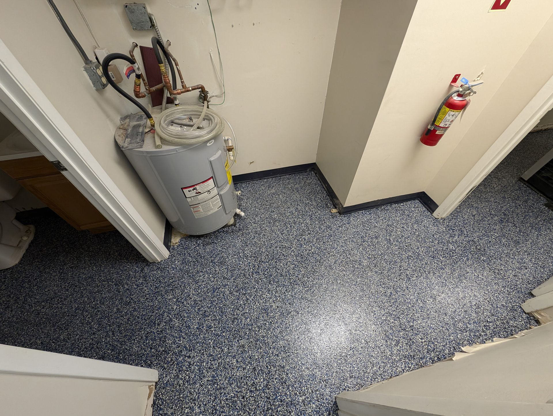 Utility closet with water heater, pipes, and a fire extinguisher on speckled gray carpet.