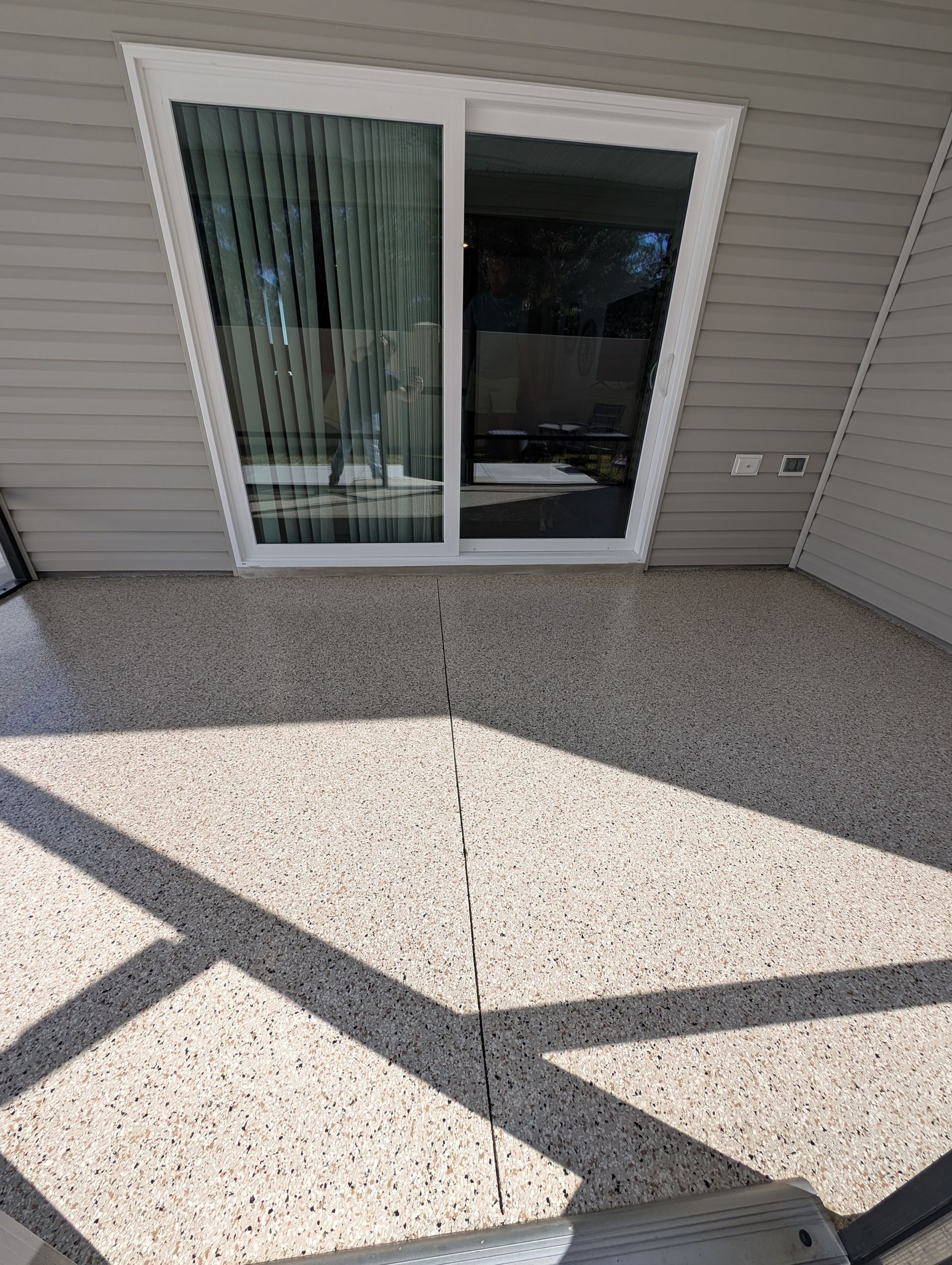 Sliding glass patio door opening to a small speckled concrete patio, with strong diagonal shadows.