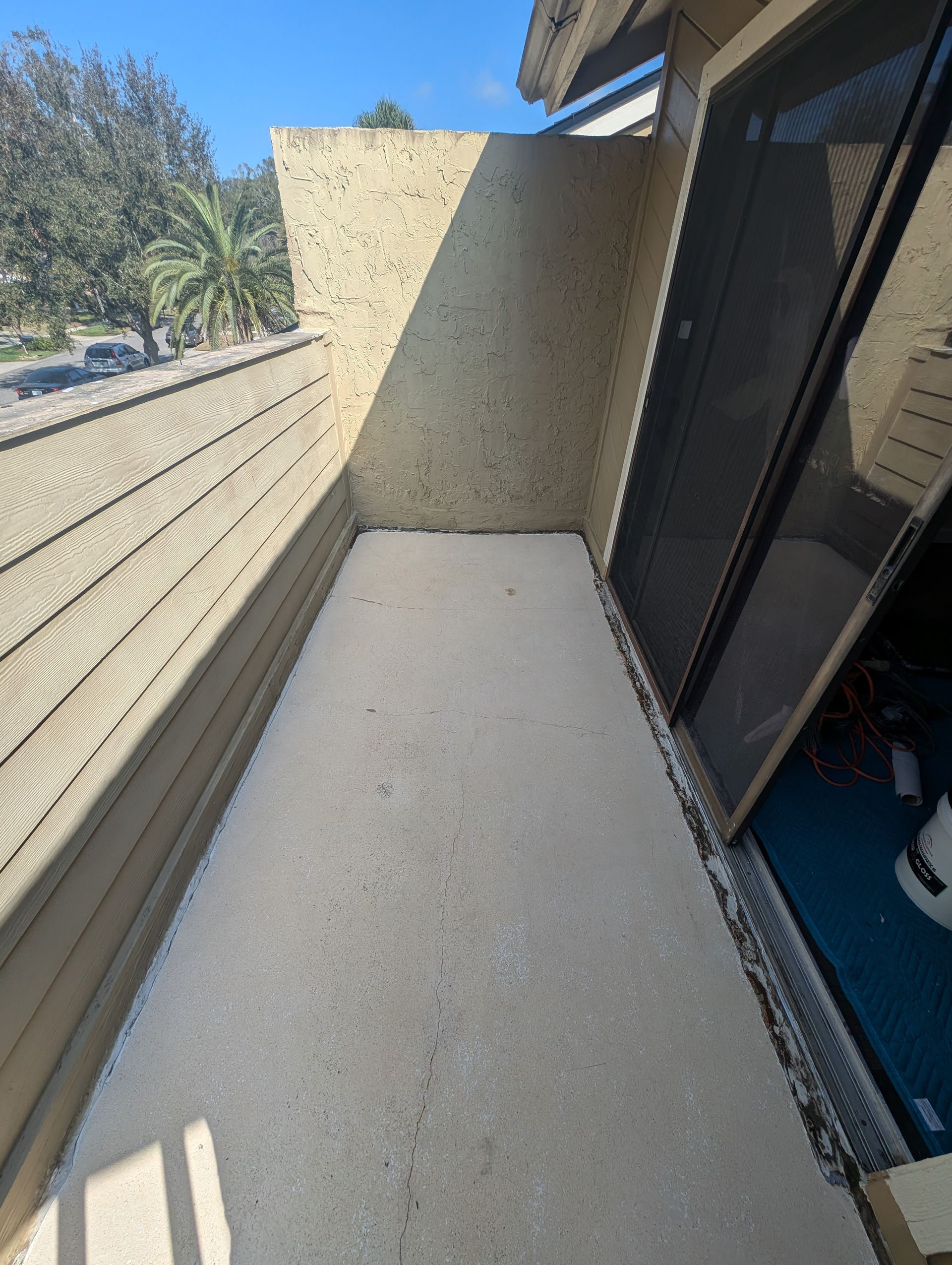Narrow concrete balcony beside a sliding glass door, with beige wall and slatted railing overlooking trees.
