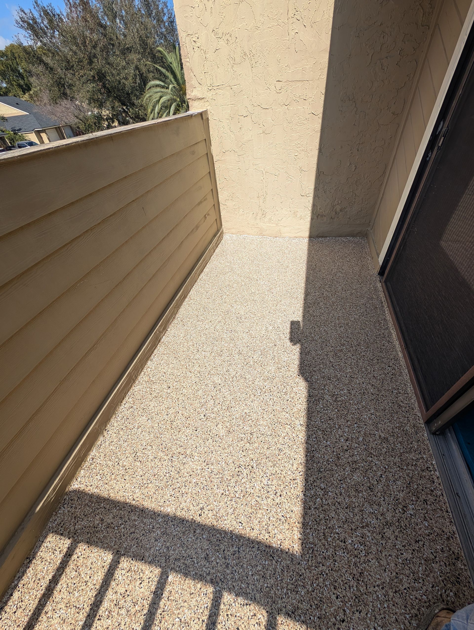 Narrow sunlit outdoor walkway with textured beige walls and a shadowed fence on the left.