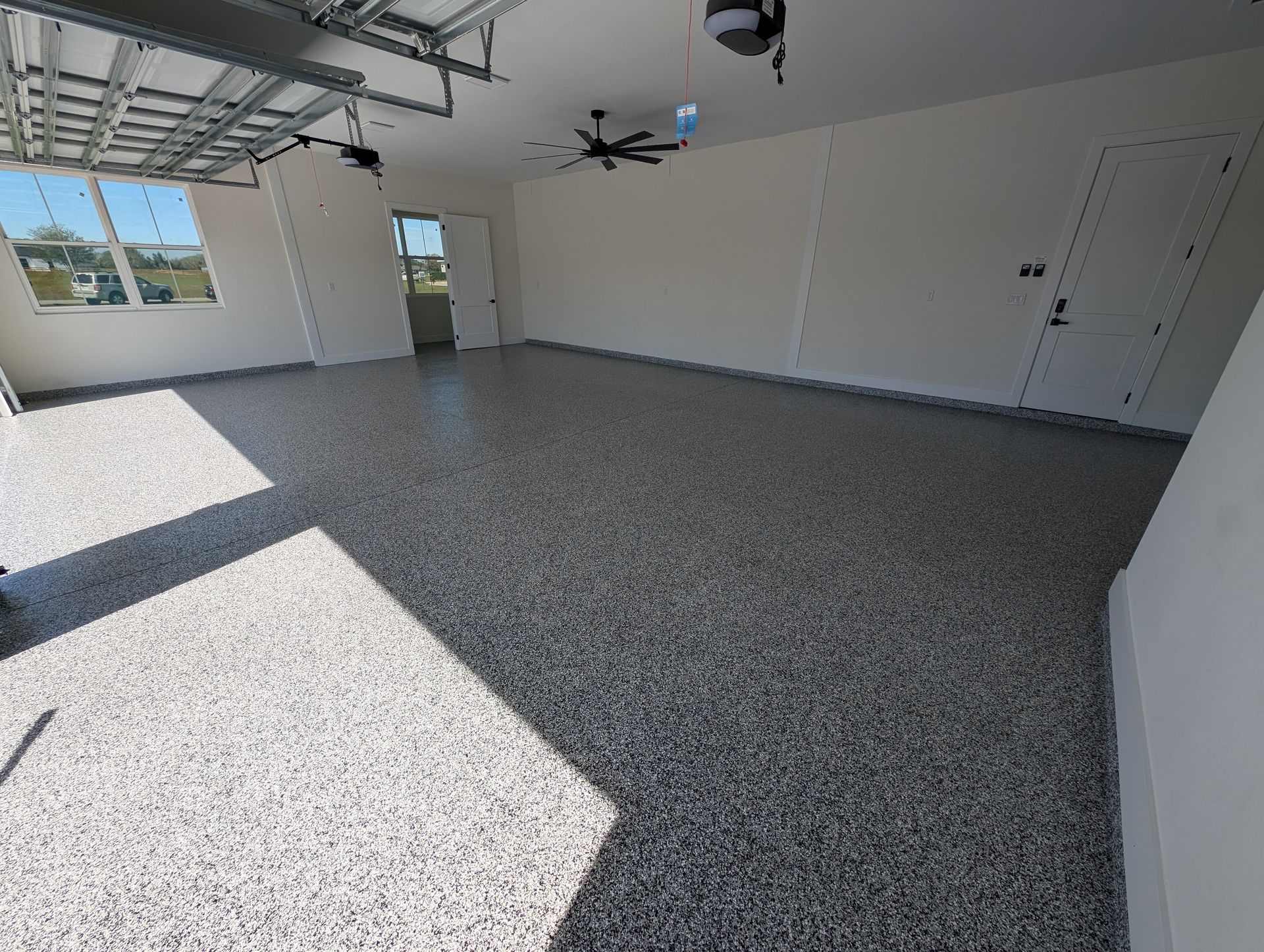Empty garage with speckled floor, white walls, large window, and overhead garage door open to sunlight