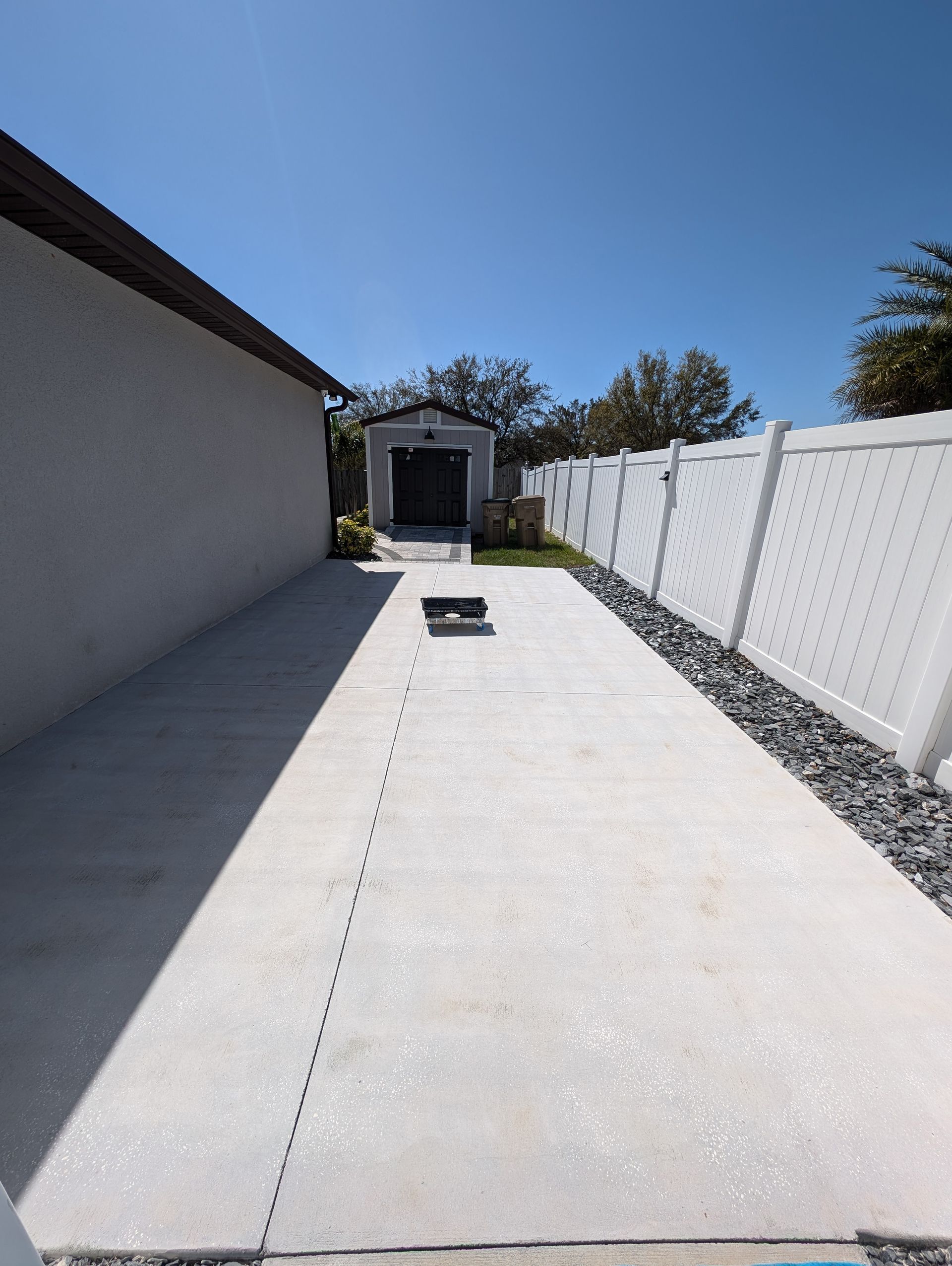 Long concrete driveway beside a white fence and house, leading to a small shed under a blue sky.