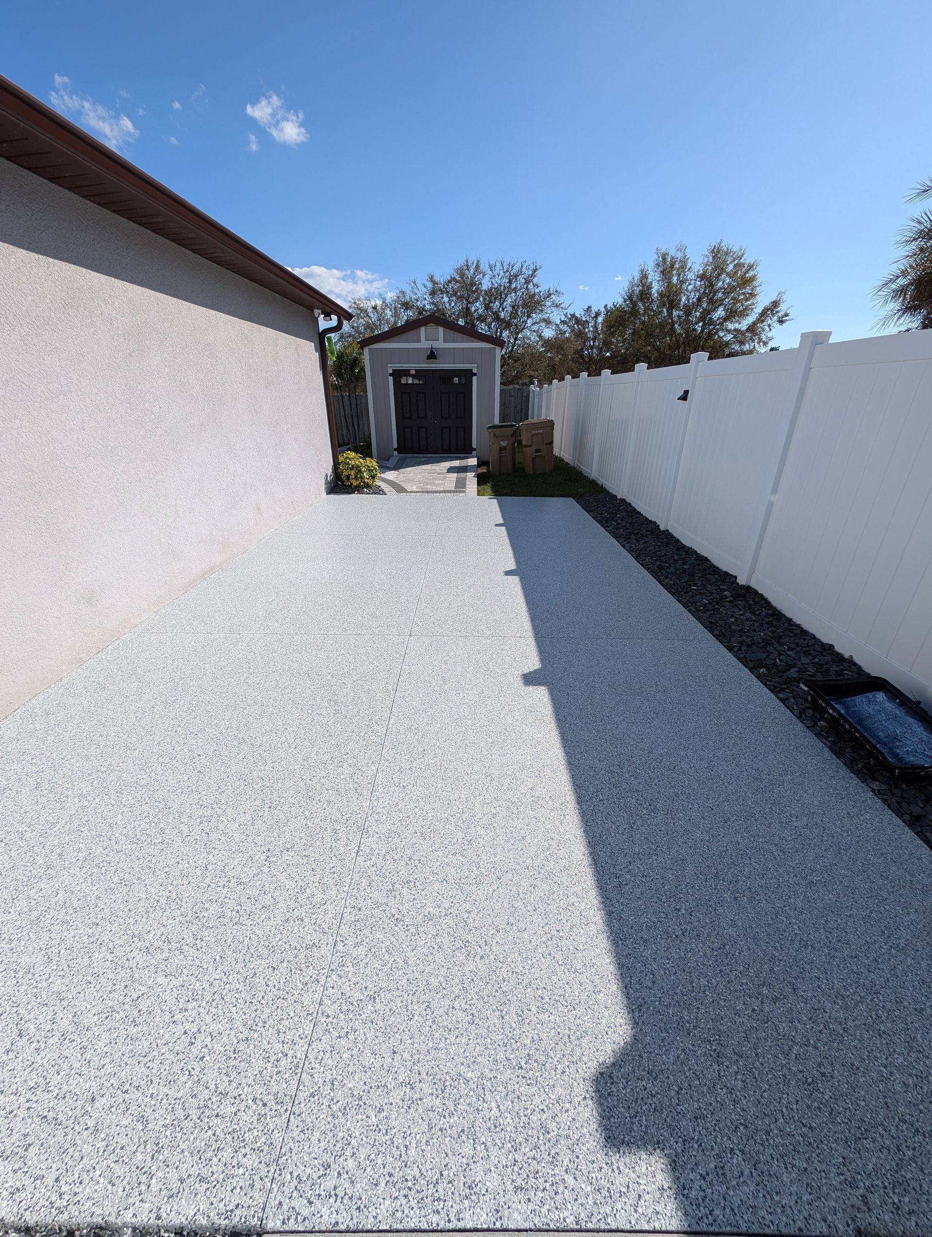 Gravel backyard path between white wall and house, leading to a small shed under a blue sky.