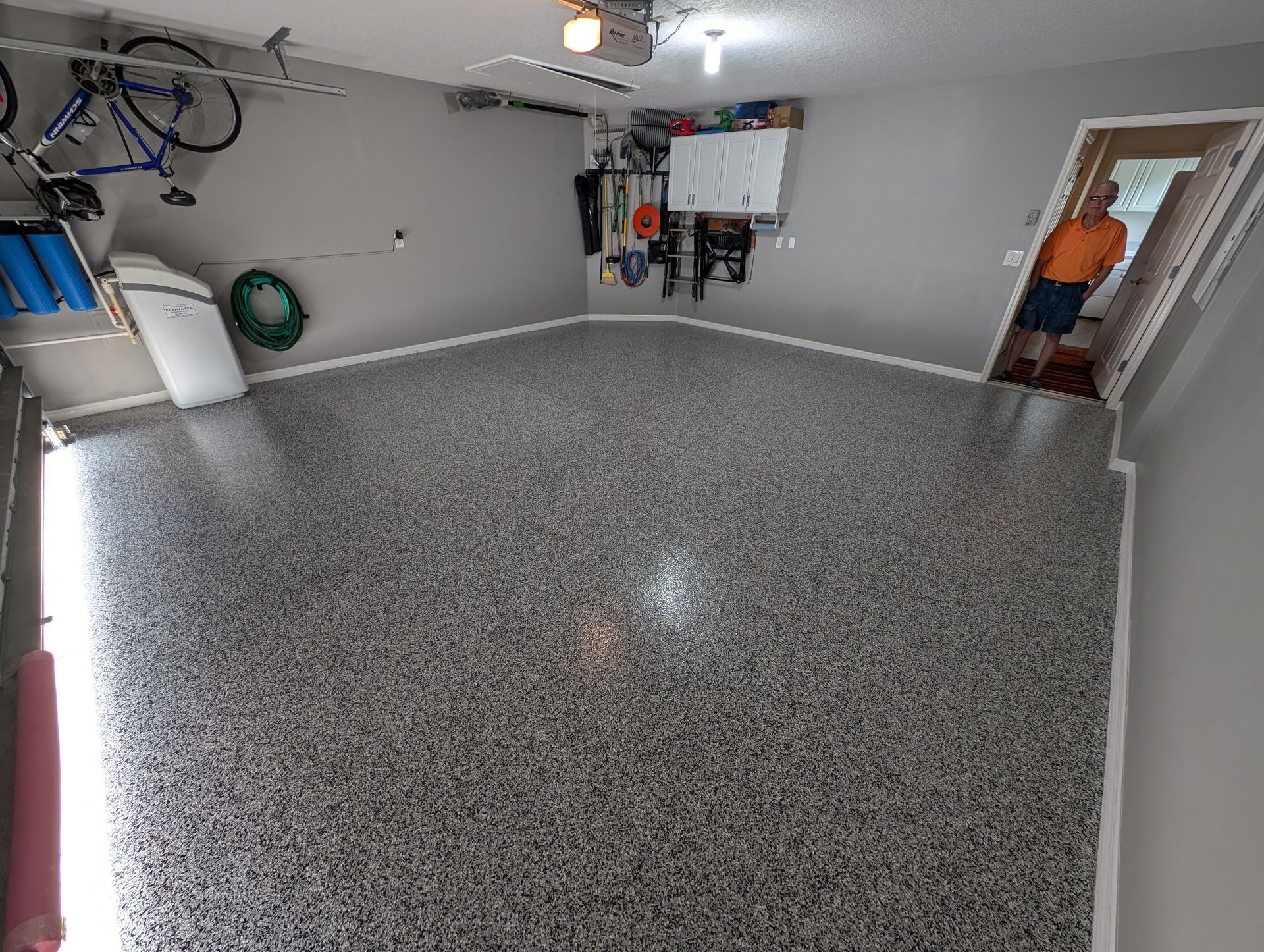 Empty garage with gray speckled floor, white walls, and water heater and tools along the back wall.