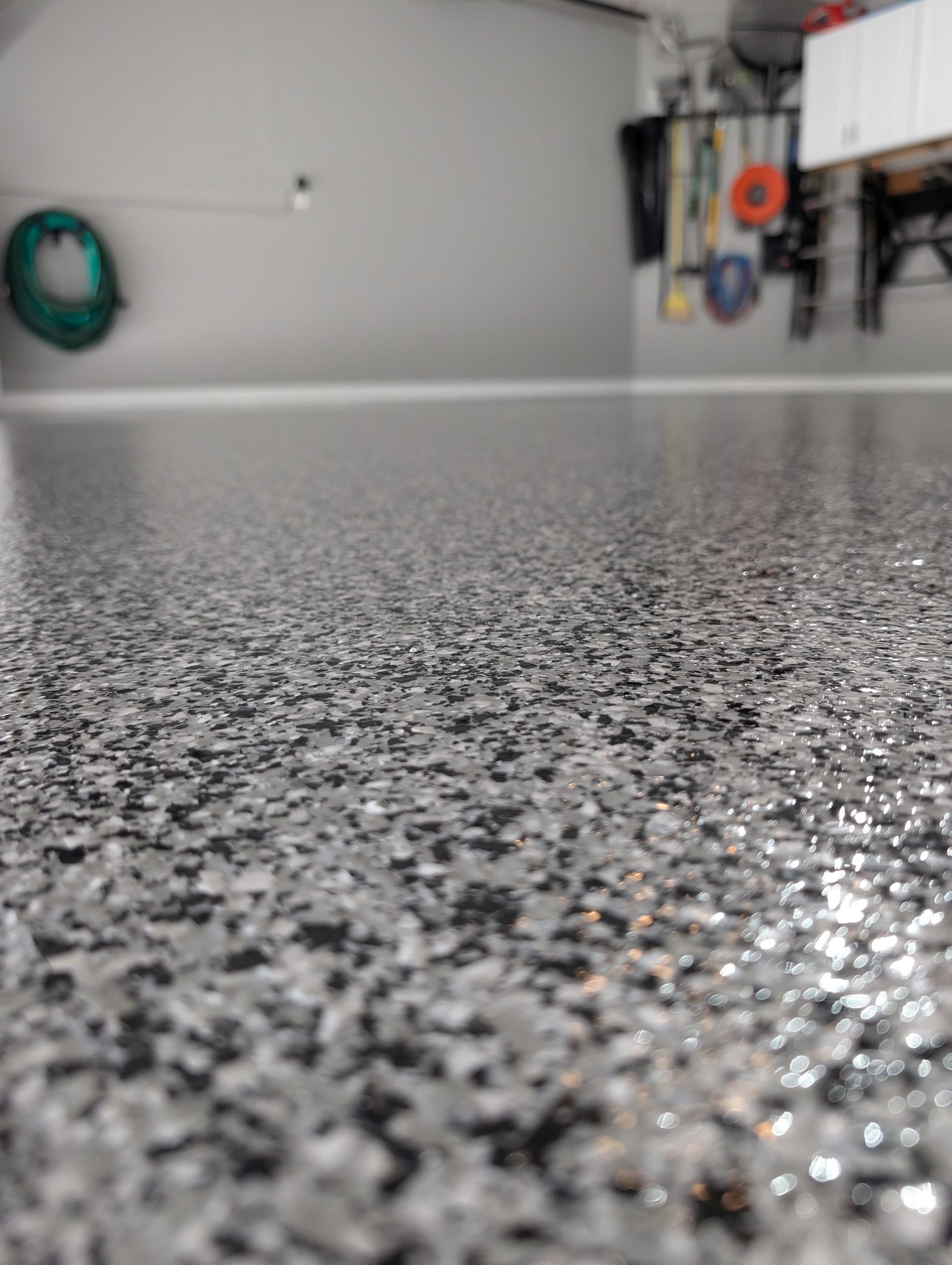 Close-up of a speckled gray epoxy garage floor with a blurred garage wall and hanging tools in the background