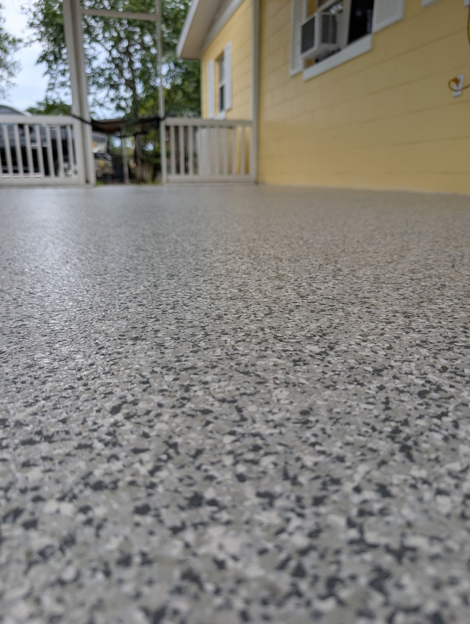Speckled gray porch floor beside a yellow house and white railing, viewed from ground level