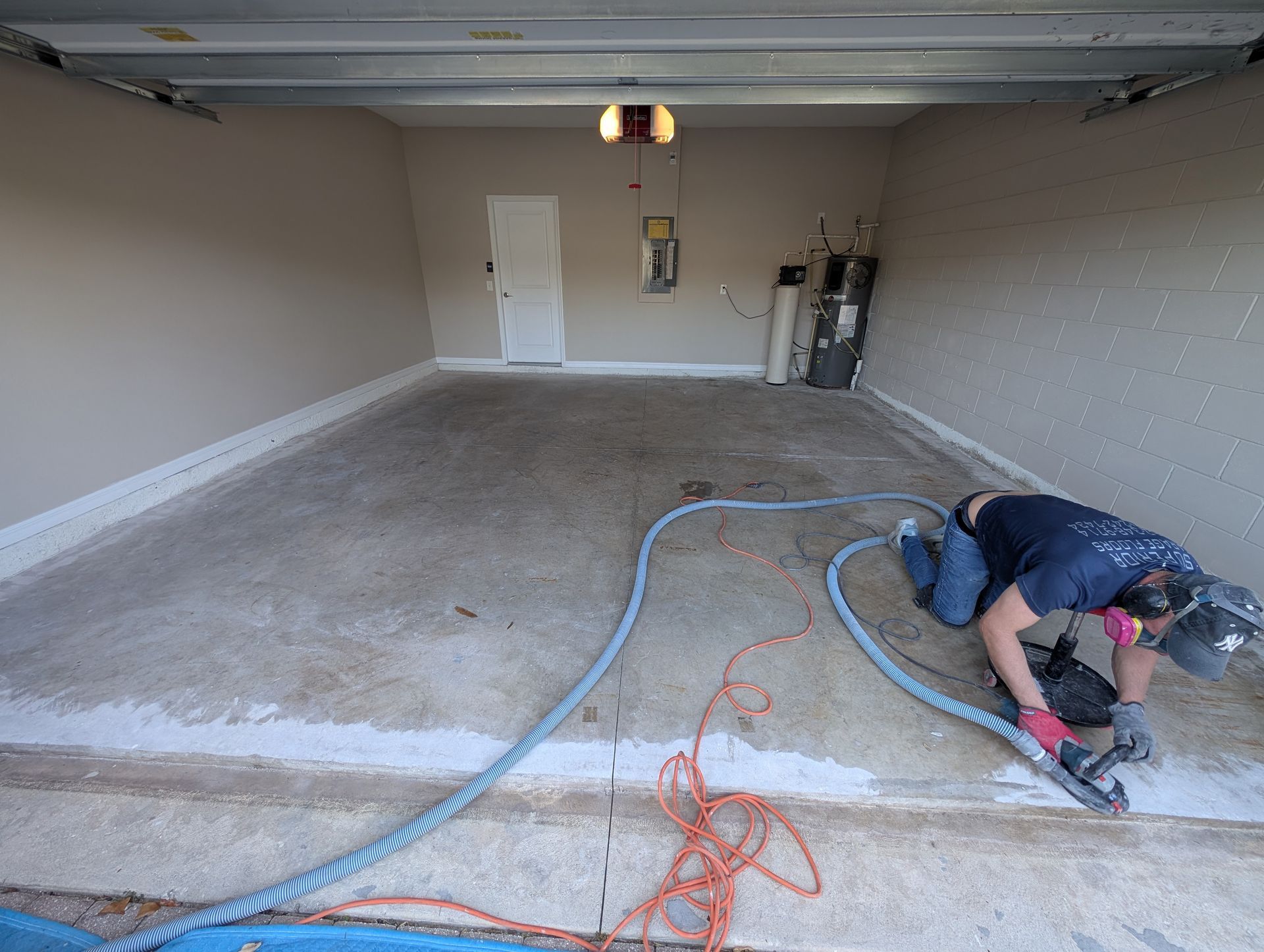 Worker sanding concrete garage floor with power tool and hoses, preparing the surface for coating.