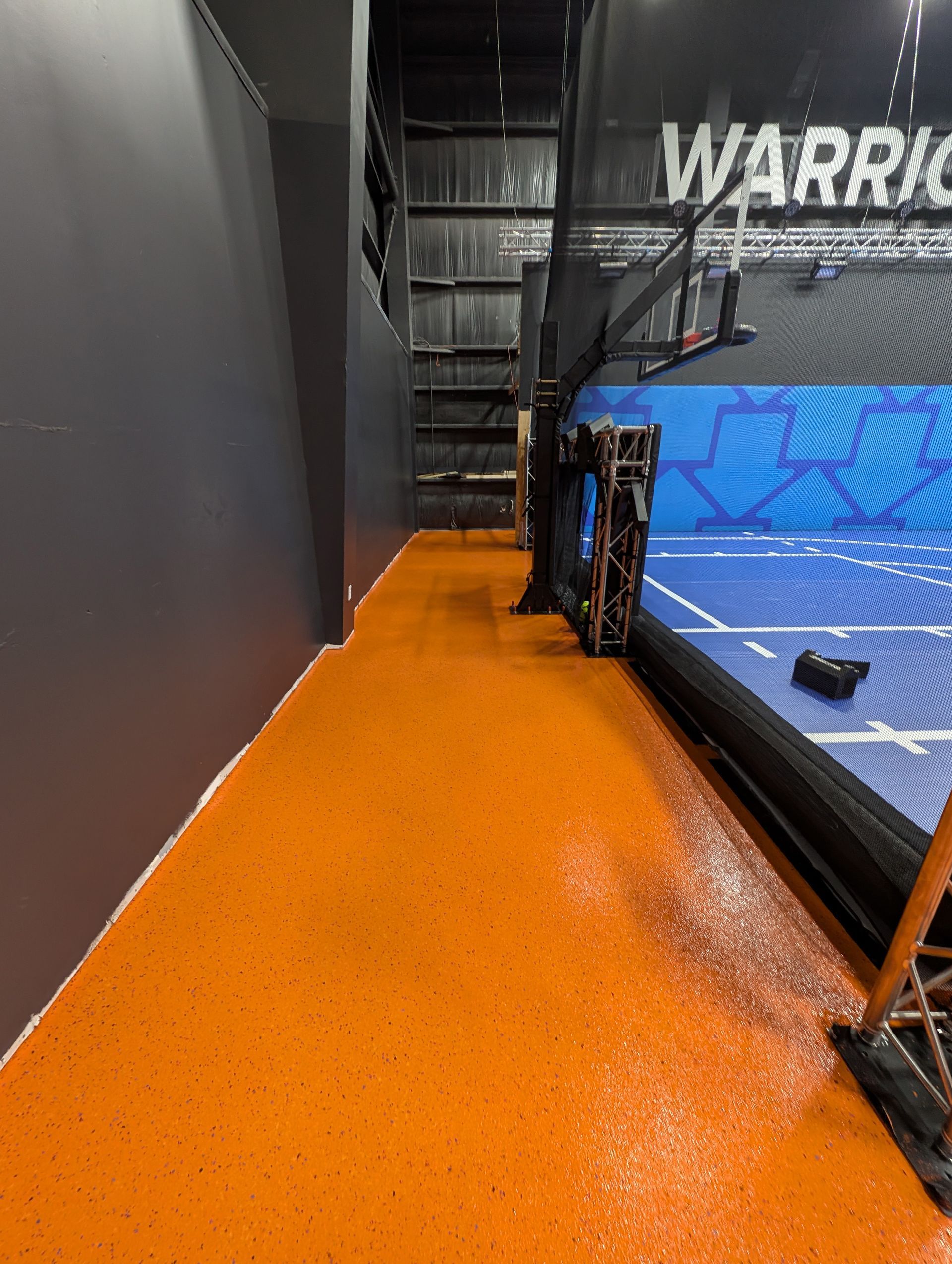 Orange padded walkway beside a martial arts ring with blue mats and black fencing