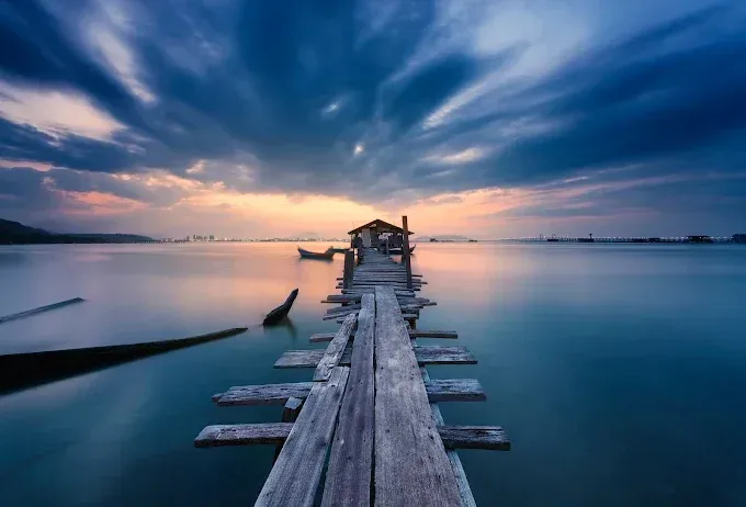 Un molo di legno si protende in un mare blu calmo al tramonto, sotto nuvole suggestive.