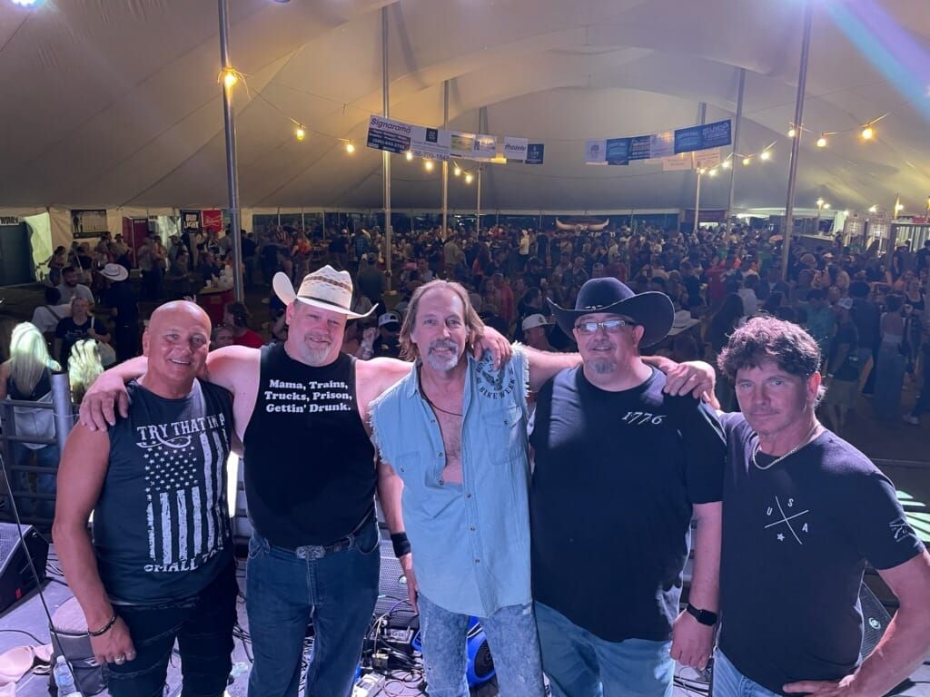 Five men posing in front of a large crowd at a nighttime concert or event.