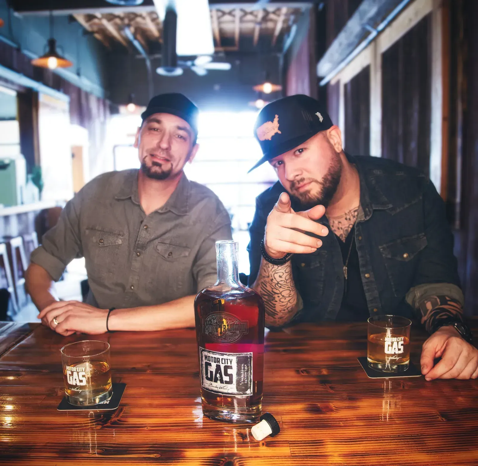Two men at a wooden bar with glasses and a bottle, one pointing at the camera in a dim pub.