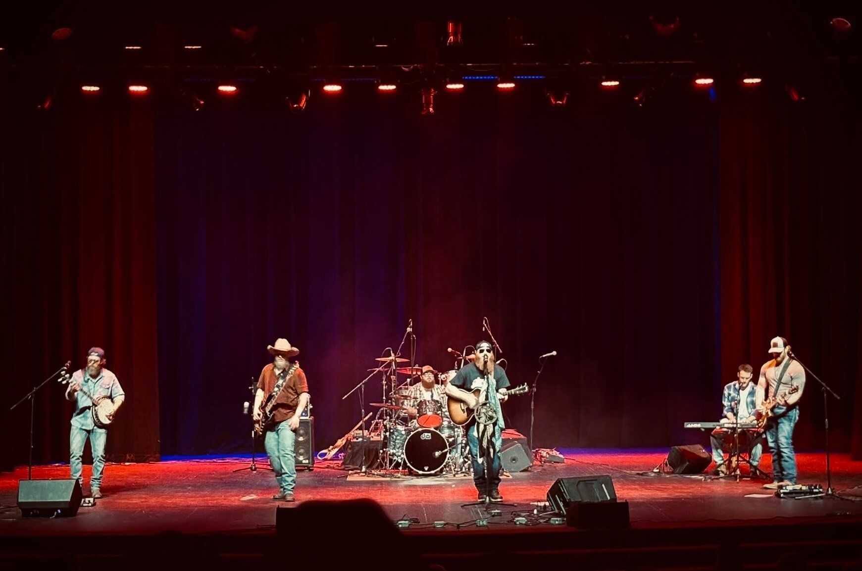 Live band performing on a red-lit stage with drums, guitars, and keyboards.