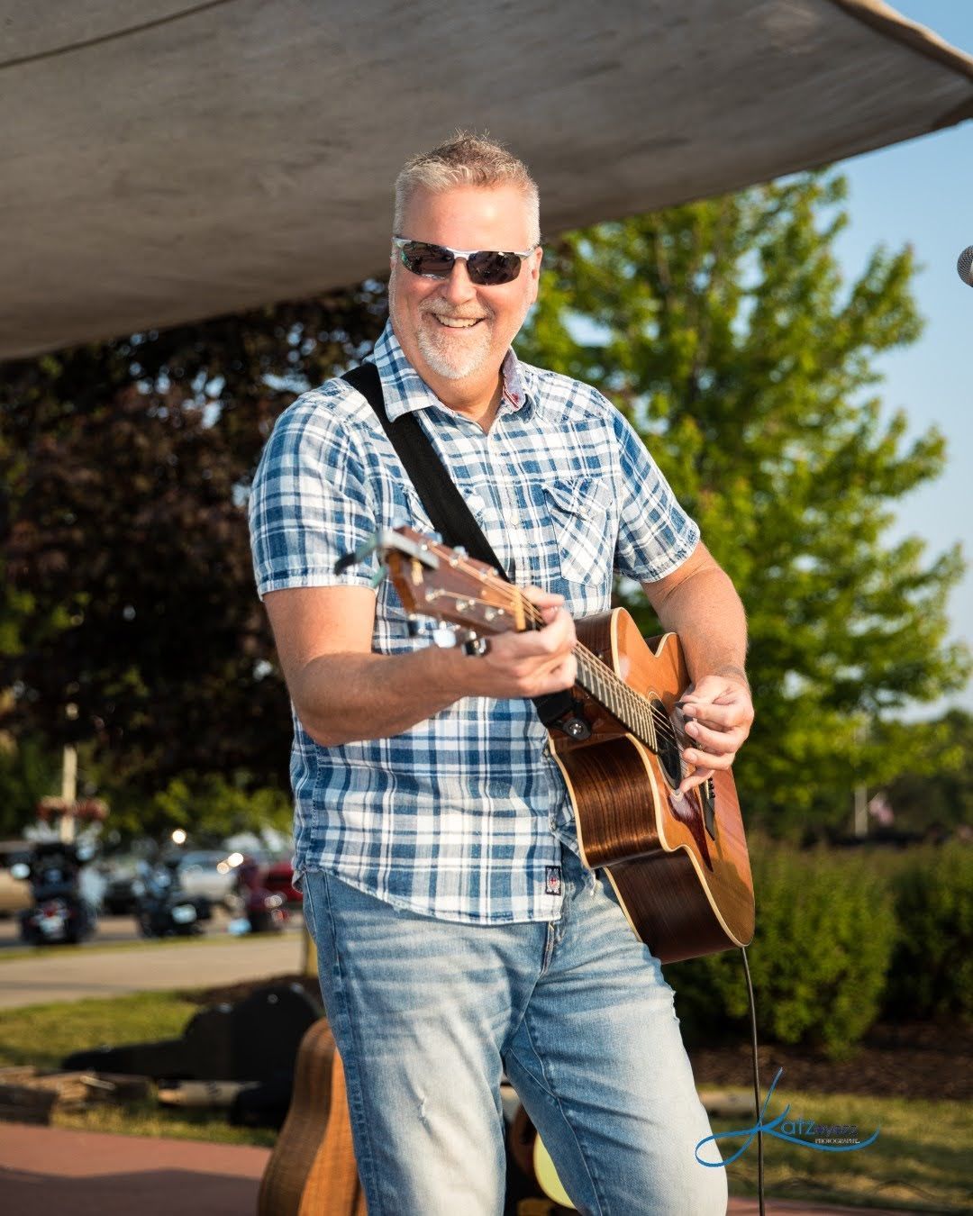 Chris Meldrum playing acoustic guitar outdoors, wearing sunglasses and a blue plaid shirt, smiling in the sun