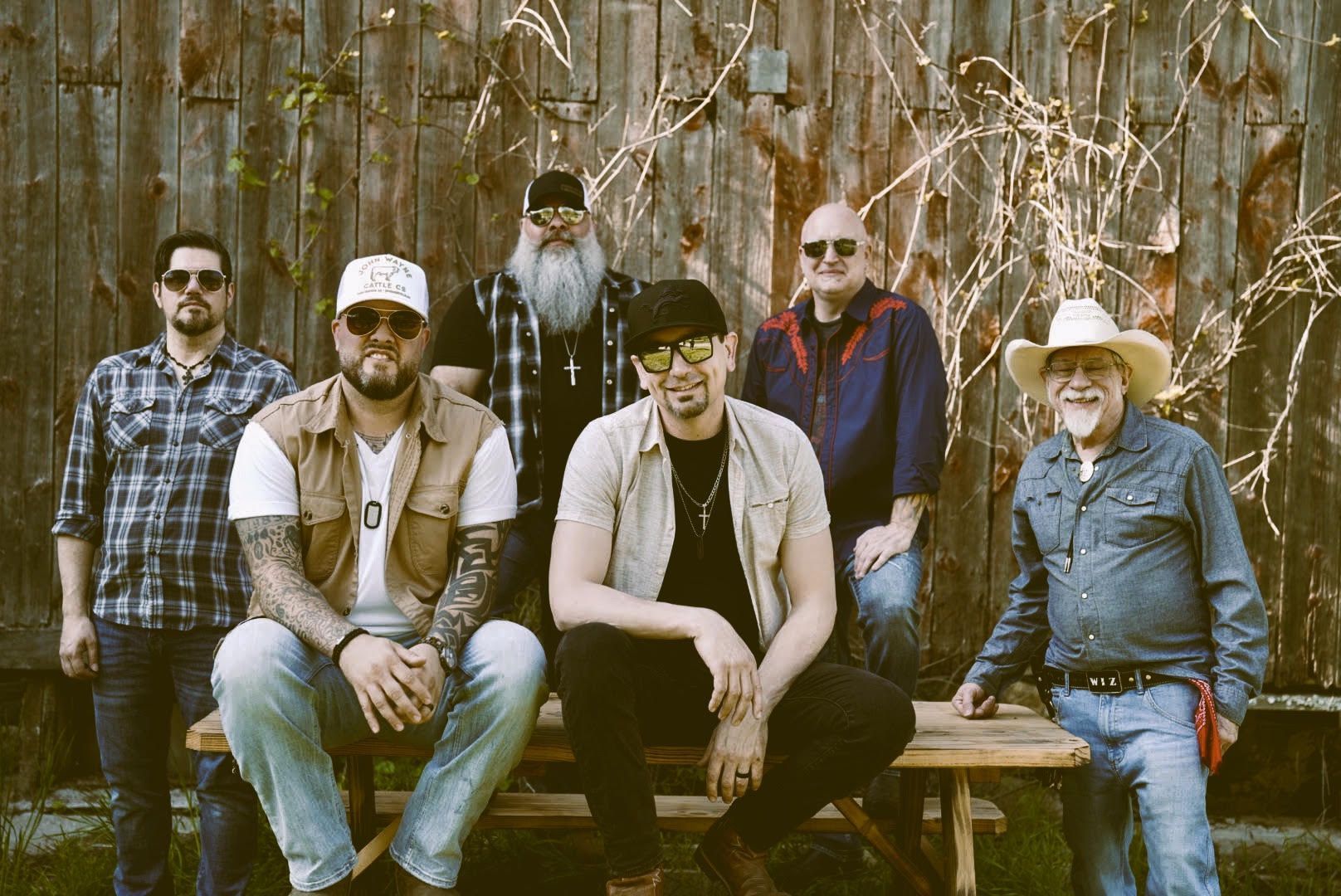 Family Tradition Band posing outdoors in front of a wooden fence and dried vines, with one in a cowboy hat.