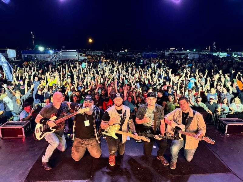 Band members posing with guitars onstage in front of a cheering crowd at night.
