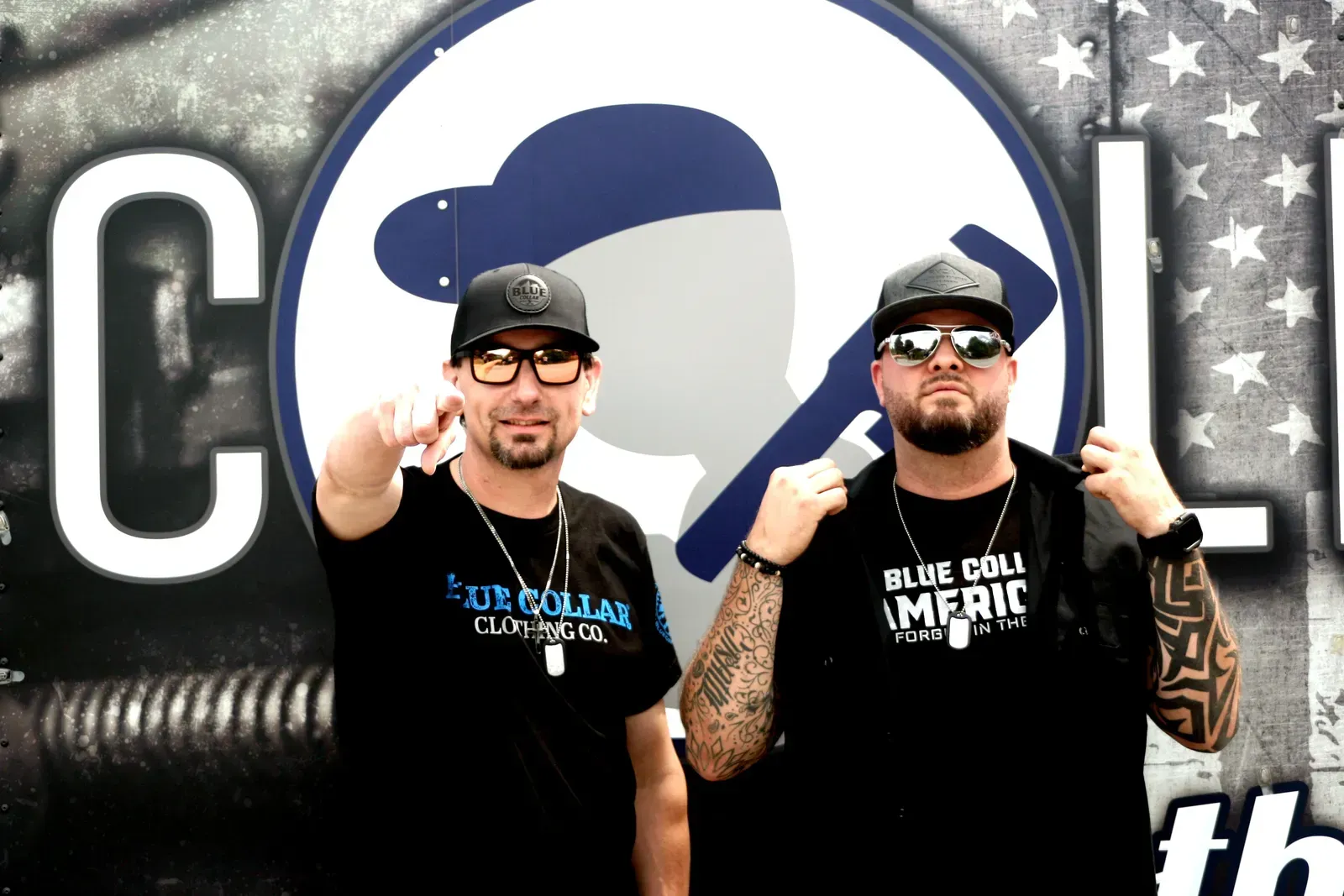 Two men in black shirts and caps pose in front of a blue-and-white logo backdrop, thumbs raised.
