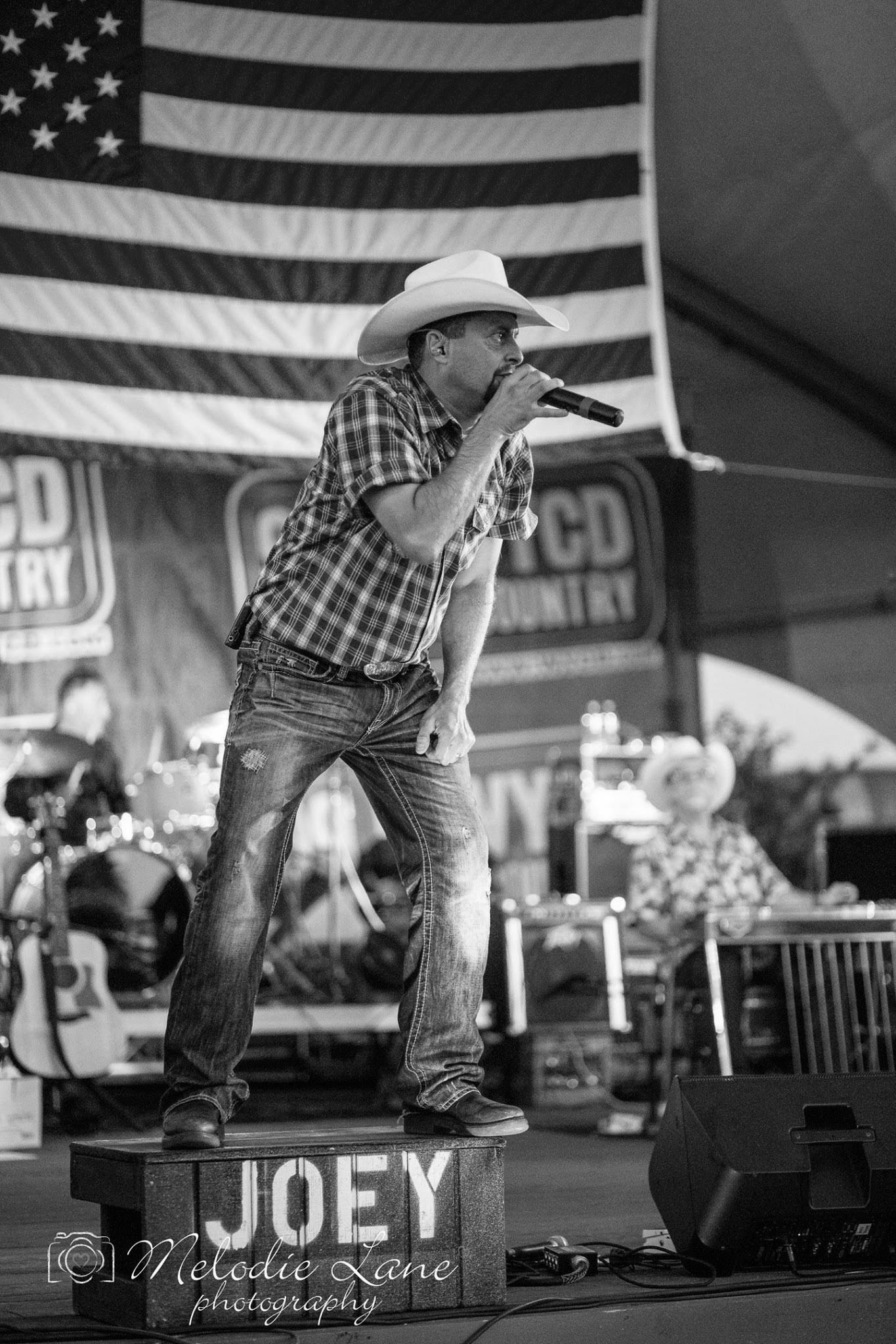 Country singer in a white hat performs on stage with a large American flag backdrop.