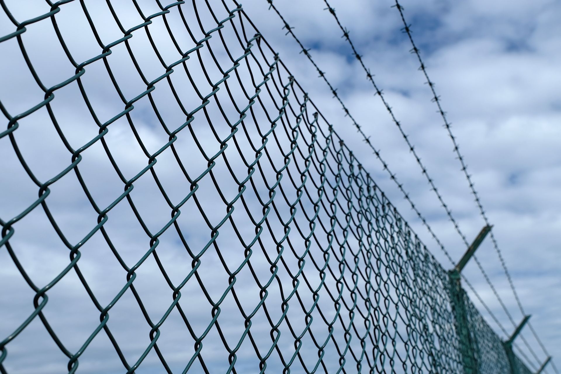 Chain-link fence topped with barbed wire against a cloudy blue sky