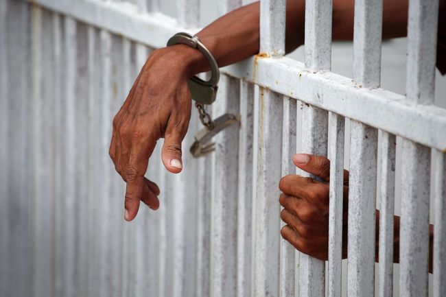 Hands gripping a white metal prison fence, one with a handcuff visible