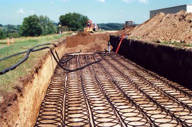 A trench in a field containing rows of coiled black geothermal piping, with heavy machinery visible in the background.