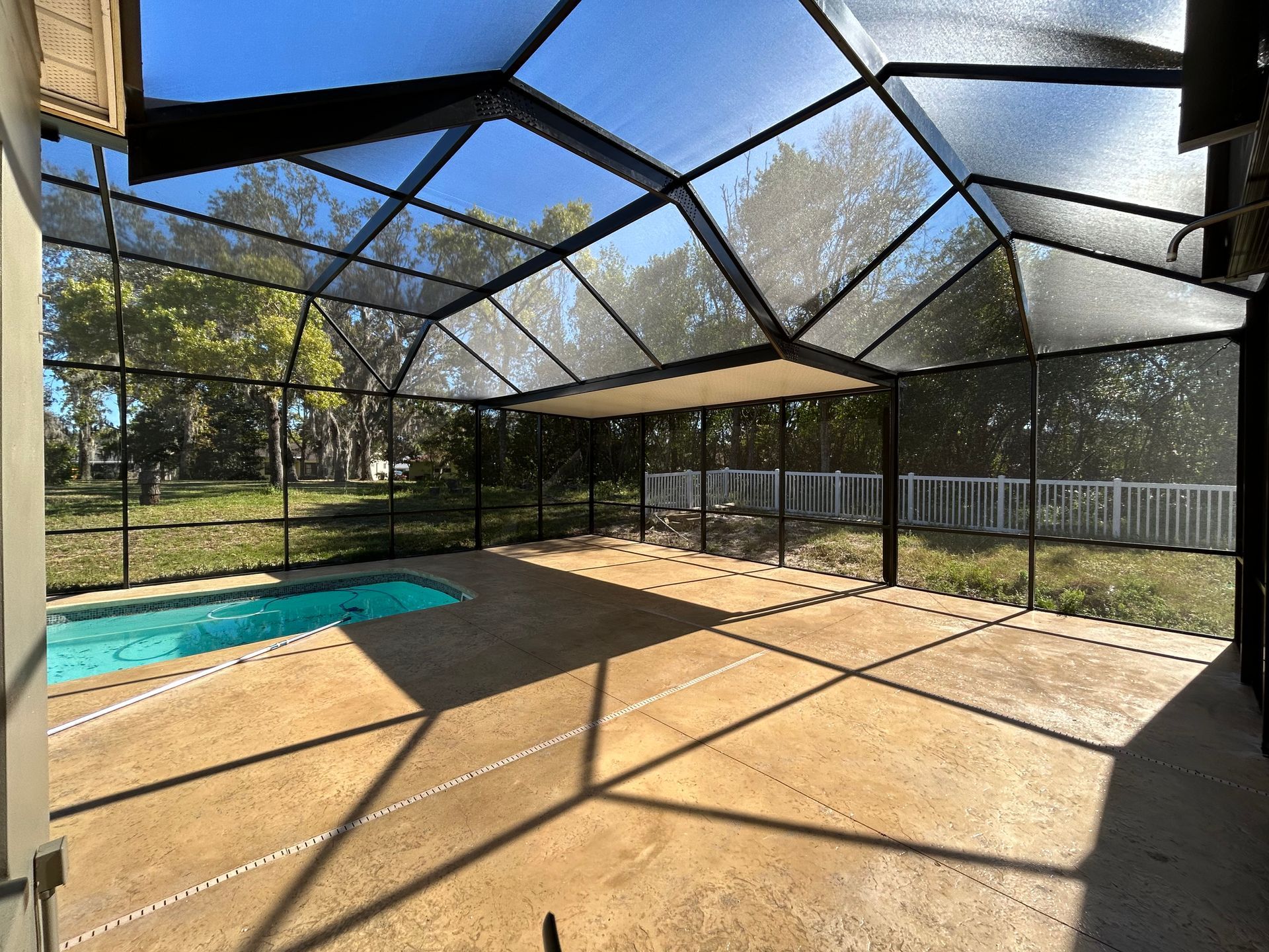 Screened-in pool enclosure with pool and patio. Black framing, beige concrete, blue sky and water, white fence, green trees.