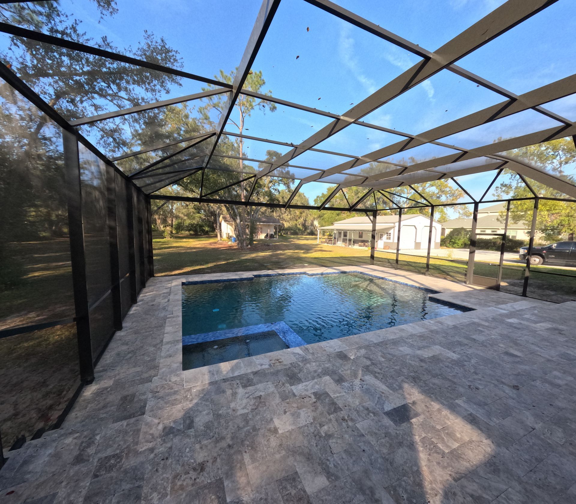 Pool under screen enclosure, stone patio, with view of backyard and houses.