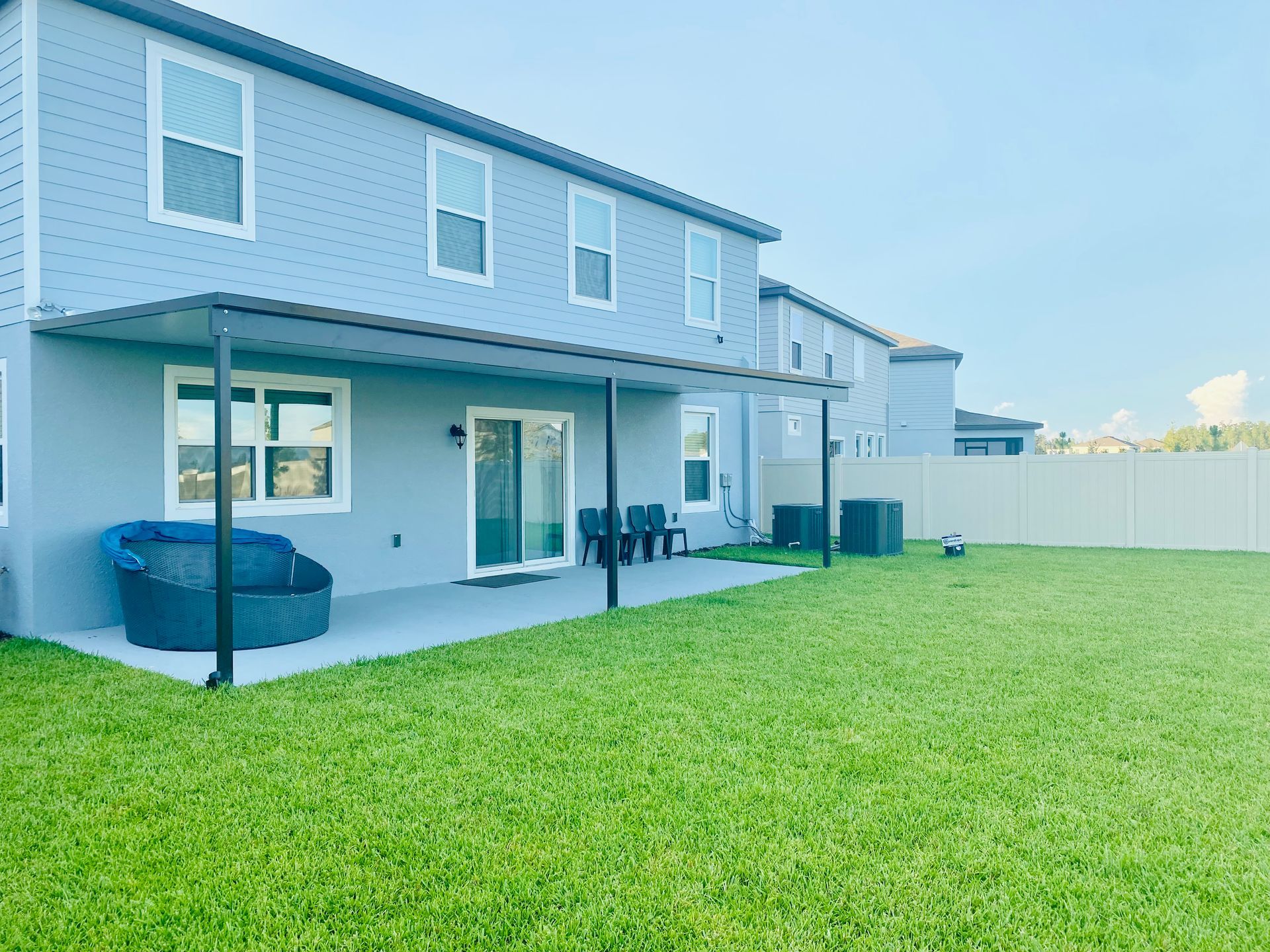 Backyard of a two-story house with patio, lawn, and fence. Blue siding, black patio cover and furniture, green grass.