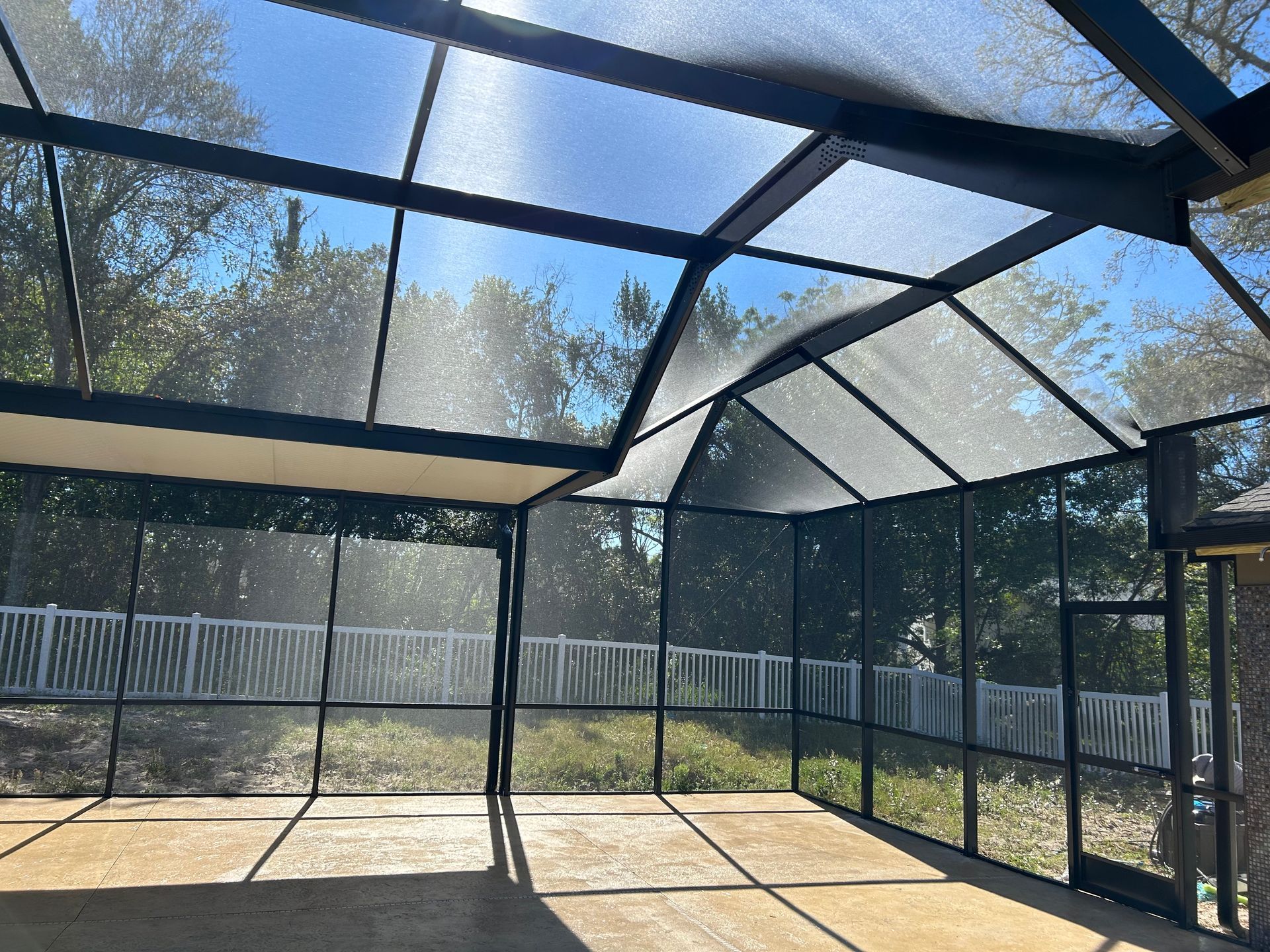 Screened-in patio with black framing and a clear, sunny sky visible. A white fence and trees are beyond.